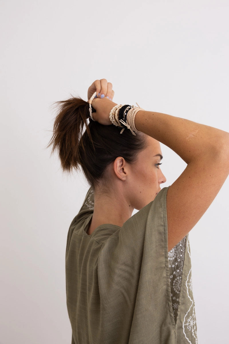 Side profile of woman with brown hair tying ponytail wearing cream multi stackable hair tie bracelets and olive green embroidered top