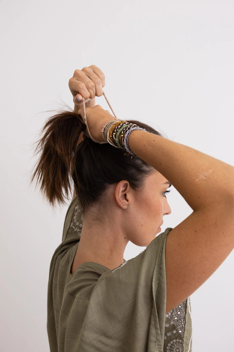 Woman with earth tone stackable hair ties and bracelets wearing olive green top tying hair back against white background