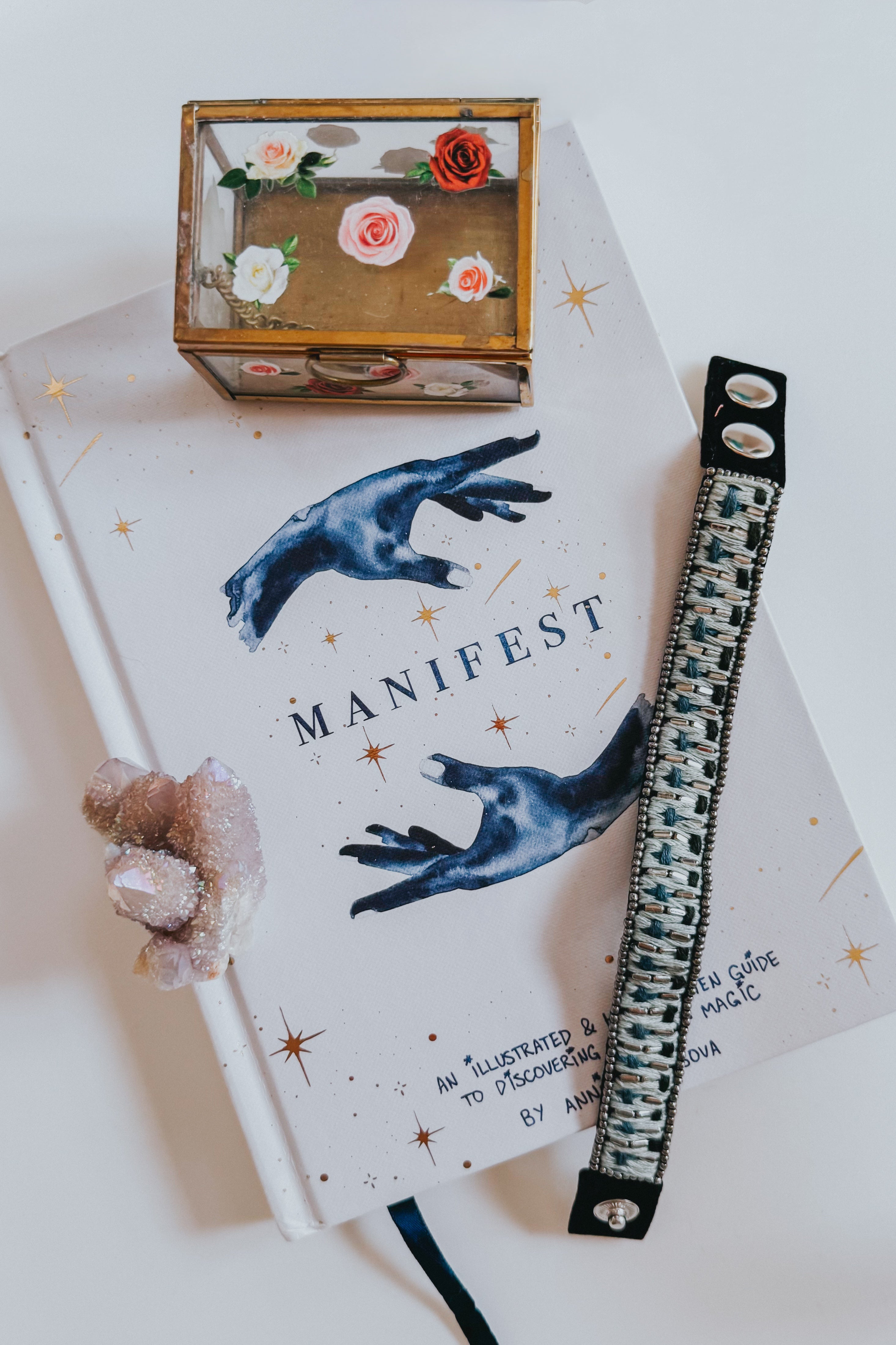 Blue woven velvet snap bracelet with intricate woven pattern resting on a white book titled Manifest with a crystal cluster and decorative box nearby