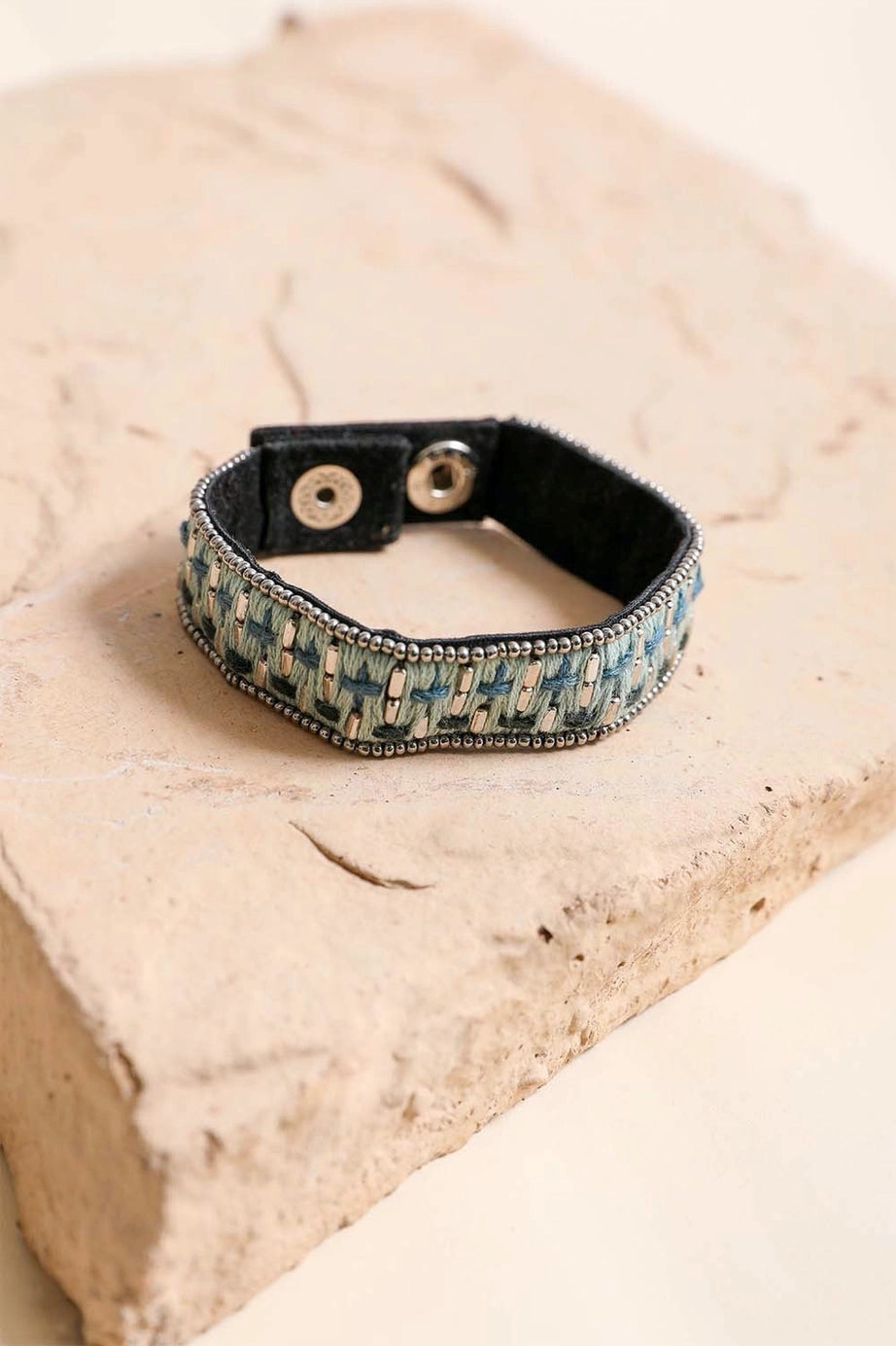 Blue woven velvet snap bracelet with silver bead trim resting on textured beige stone surface