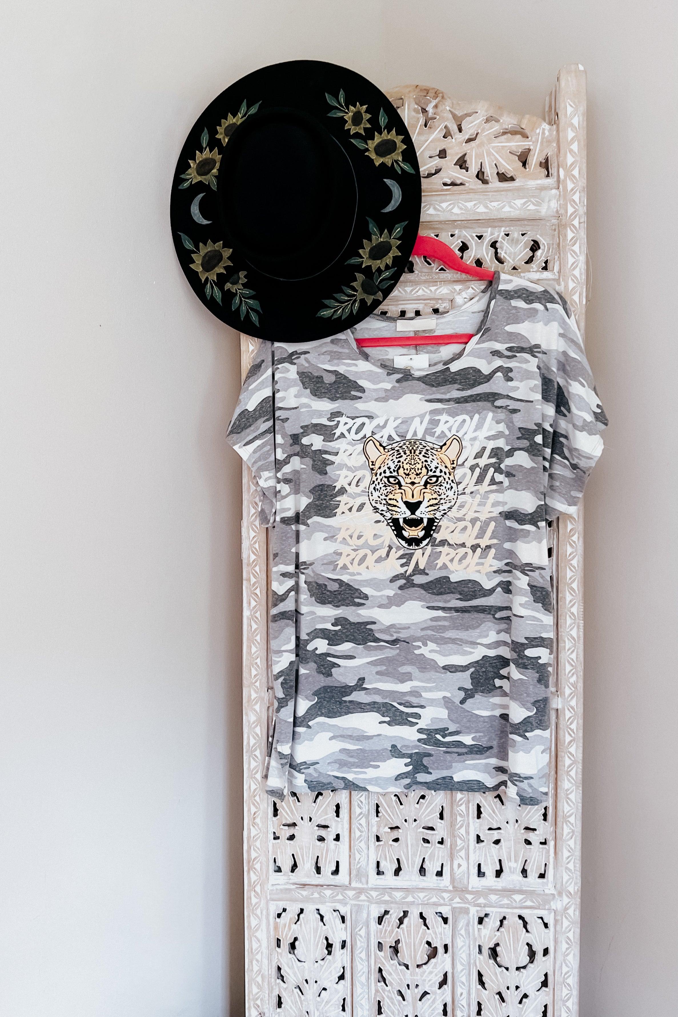 Grey camo tee with roaring leopard graphic and repeated text hanging on intricate carved wooden panel with black floral hat