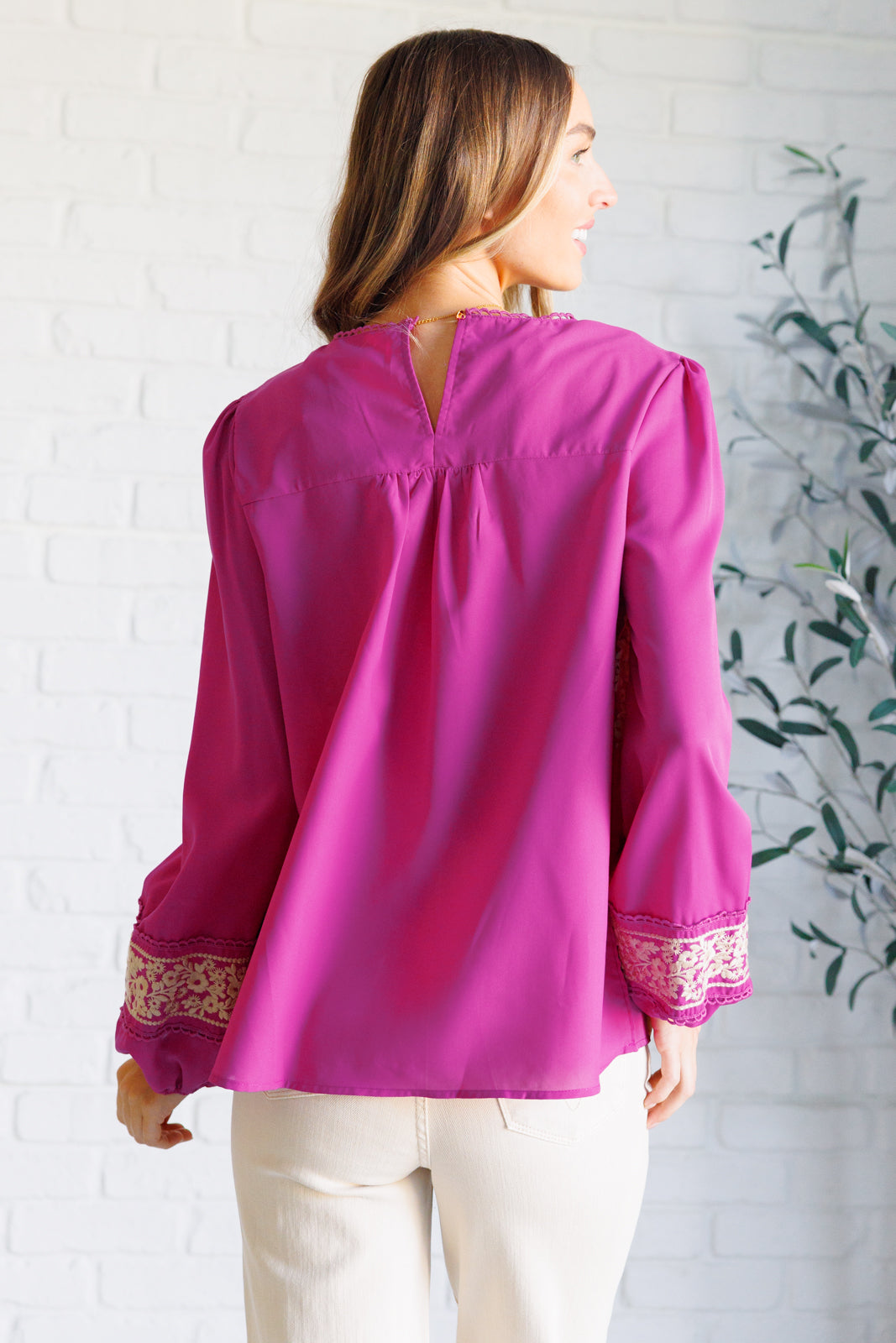 Woman wearing a magenta long-sleeve blouse with intricate white embroidery and beige jeans standing against a white brick wall.