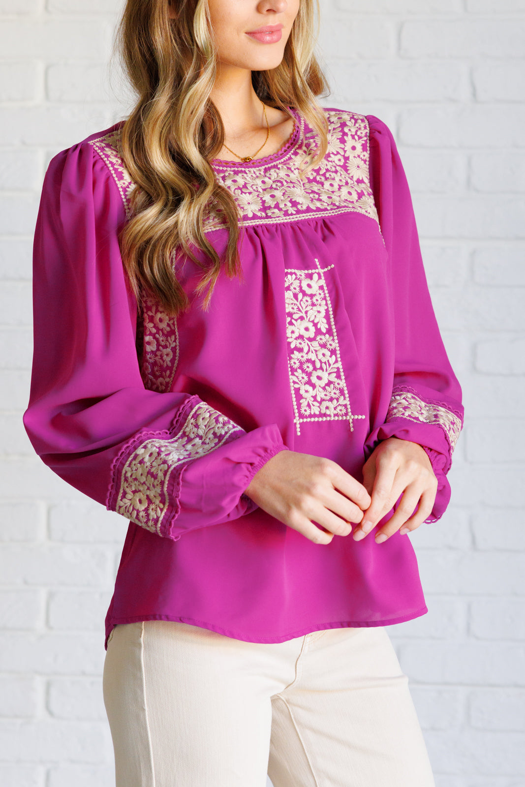 Woman wearing a magenta long-sleeve blouse with intricate white embroidery and beige jeans standing against a white brick wall.