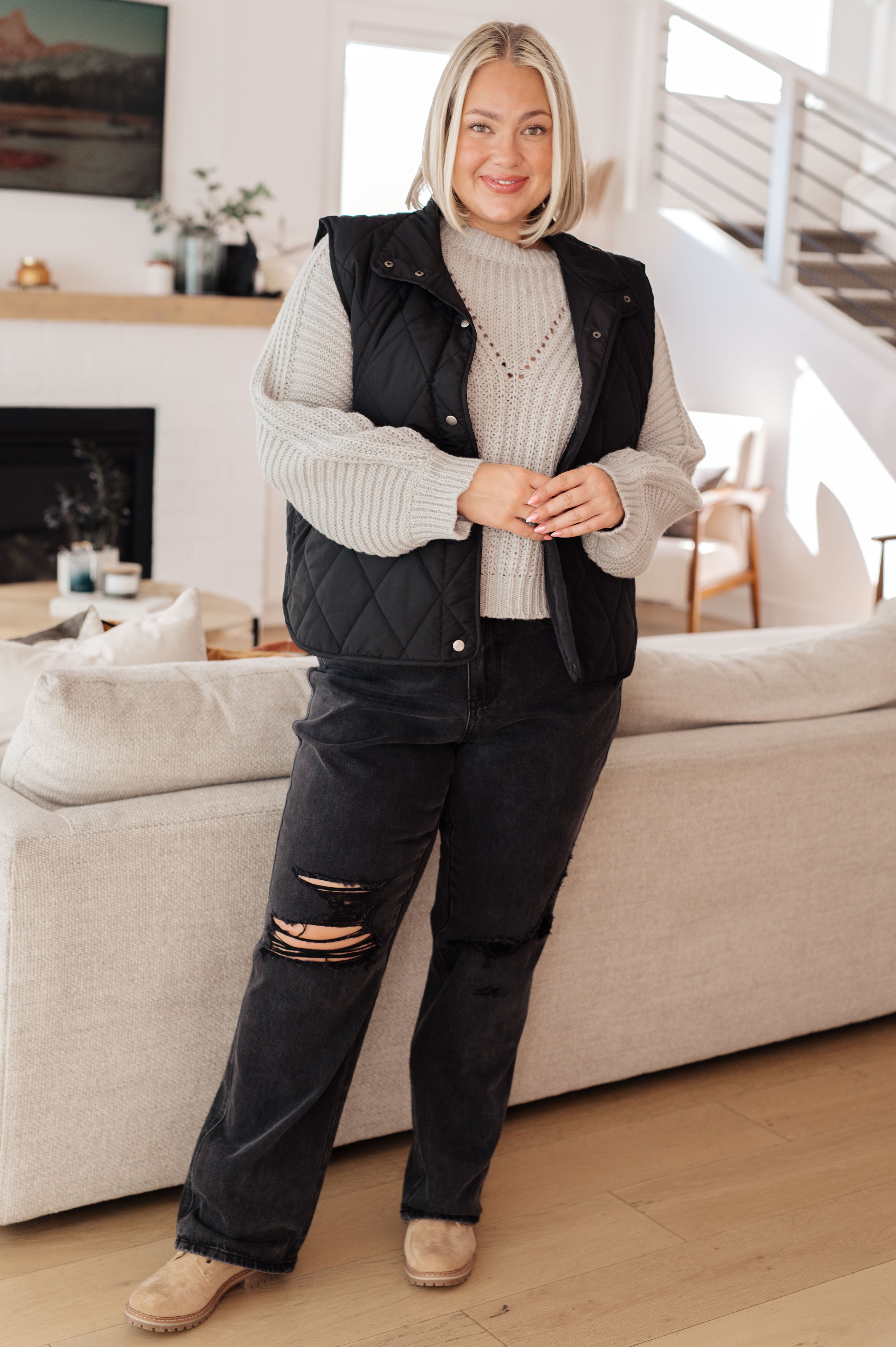 Smiling woman wearing grey balloon sleeve sweater under black quilted vest with distressed black jeans and beige shoes indoors