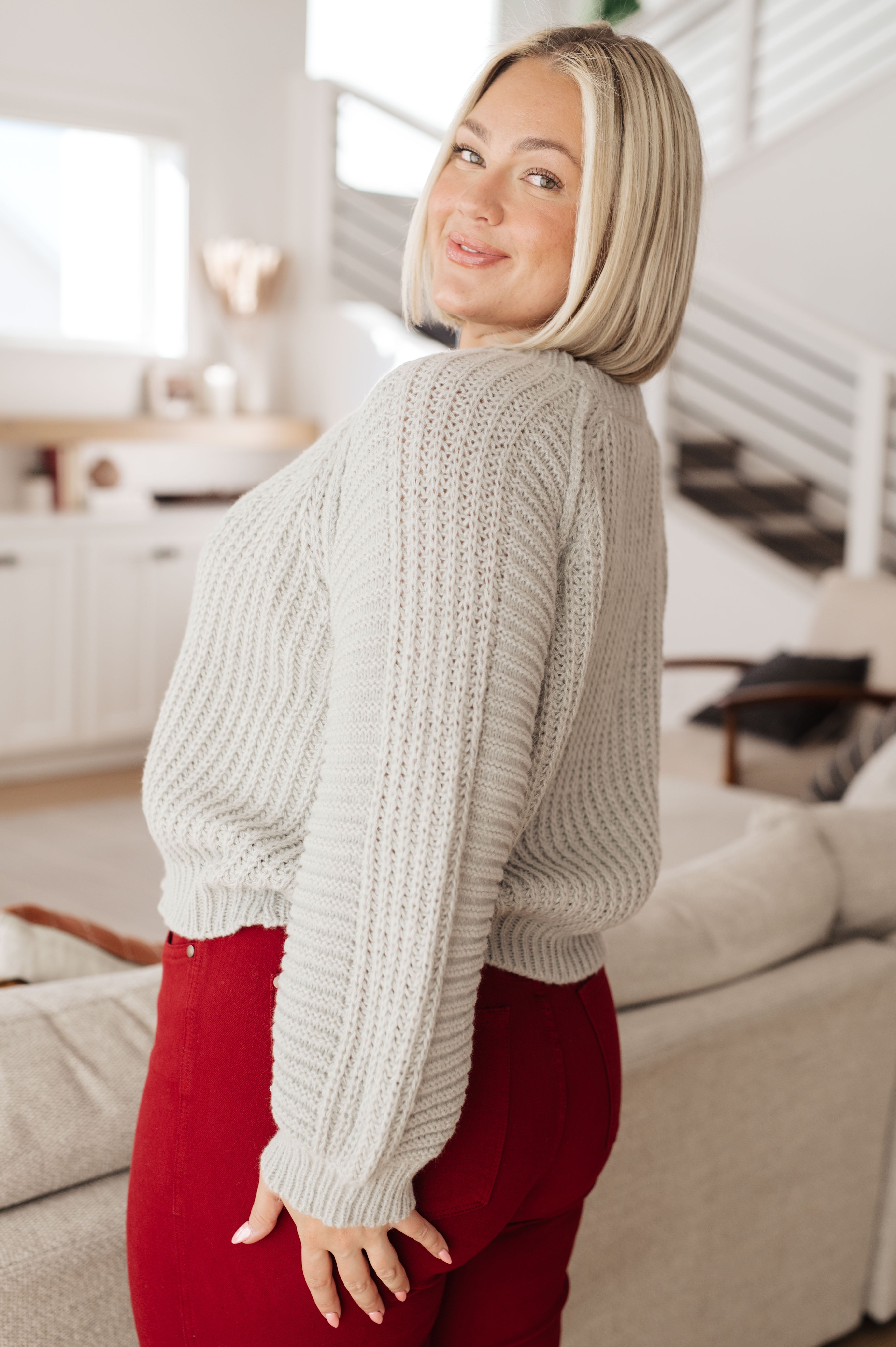Woman smiling wearing light grey balloon sleeve sweater and red pants in bright modern living room