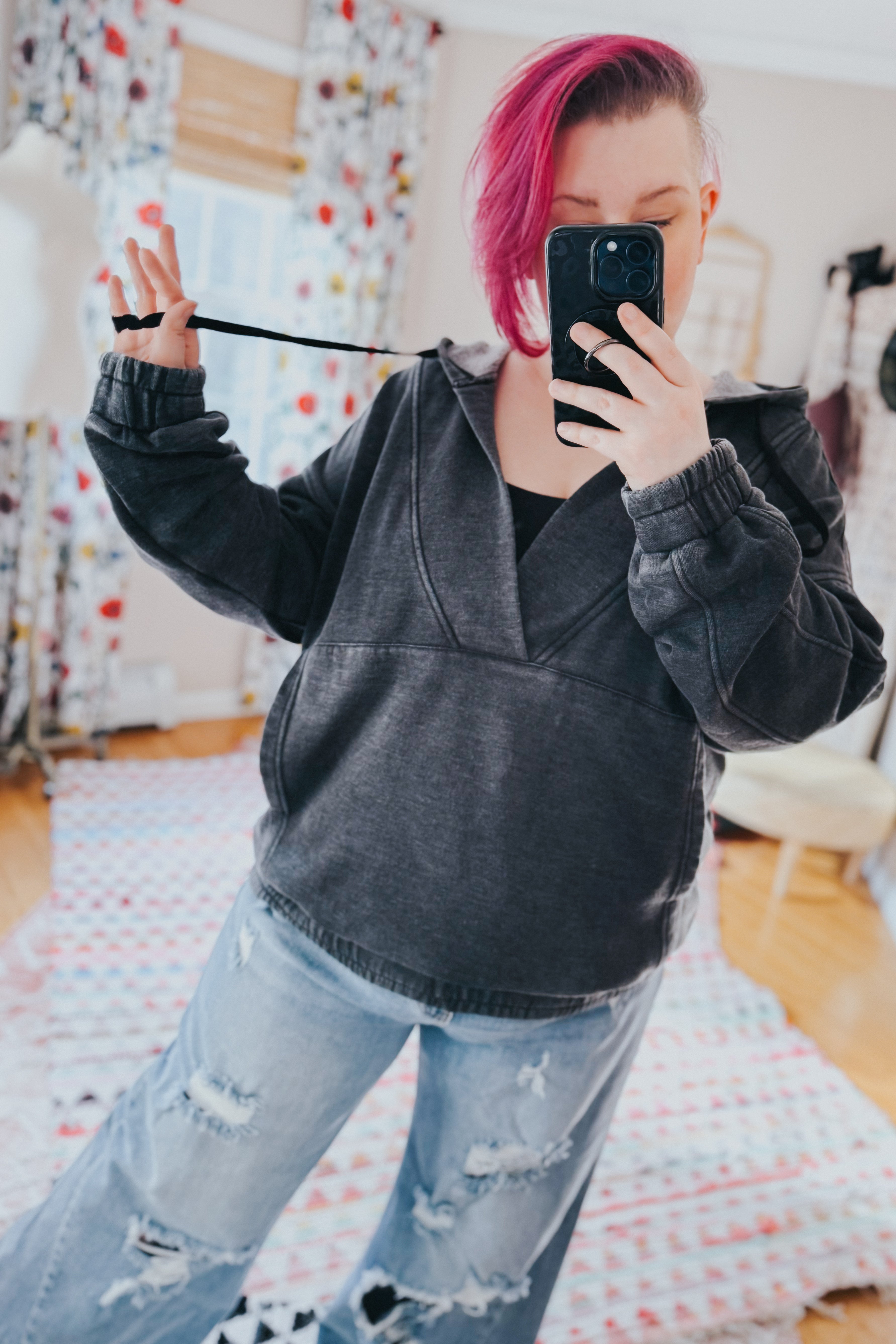 Woman with pink hair taking mirror selfie wearing charcoal acid wash hoodie and distressed denim jeans in bright room
