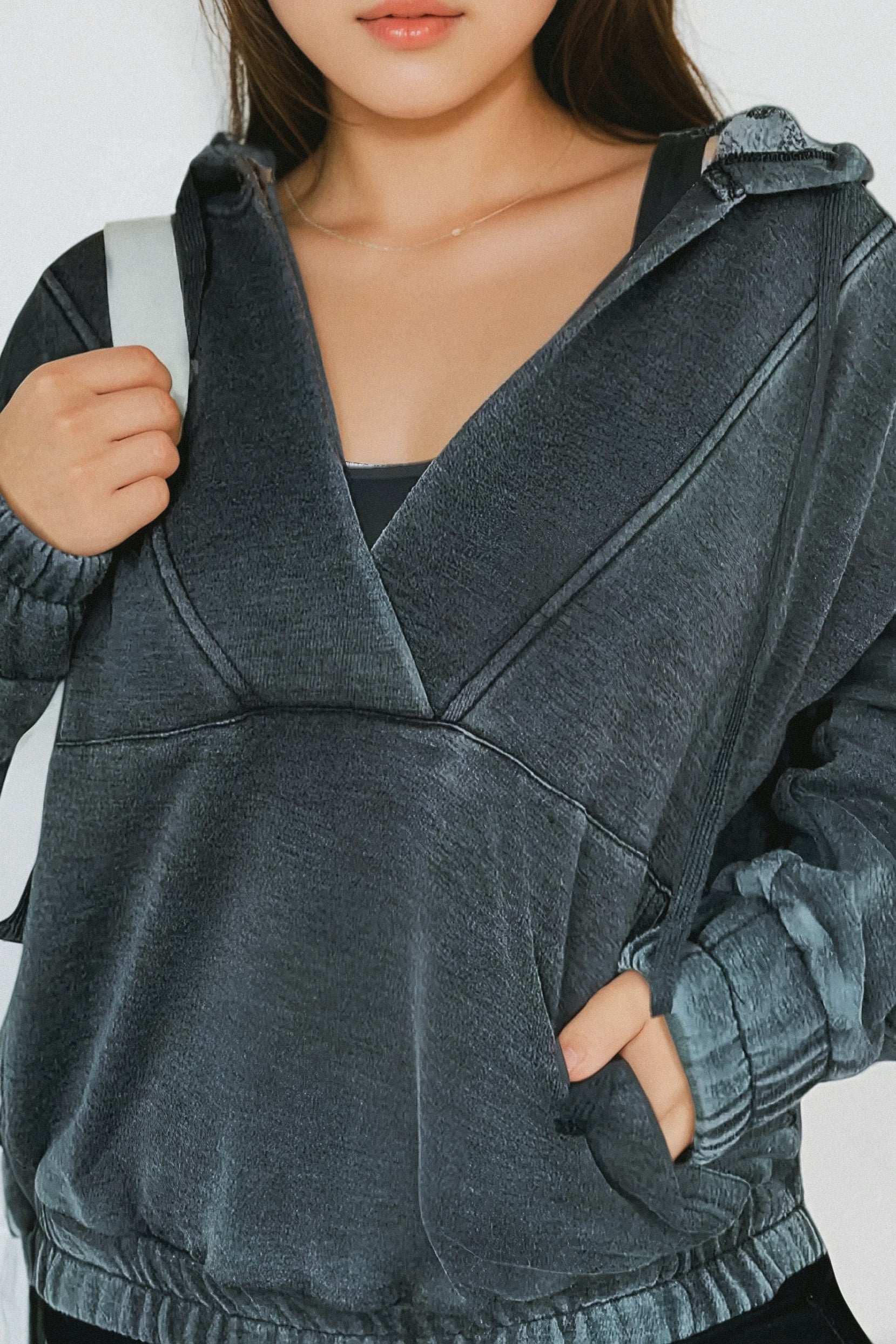 Close-up of a woman wearing a charcoal acid wash hoodie with gathered cuffs and a front pocket, holding a bag strap