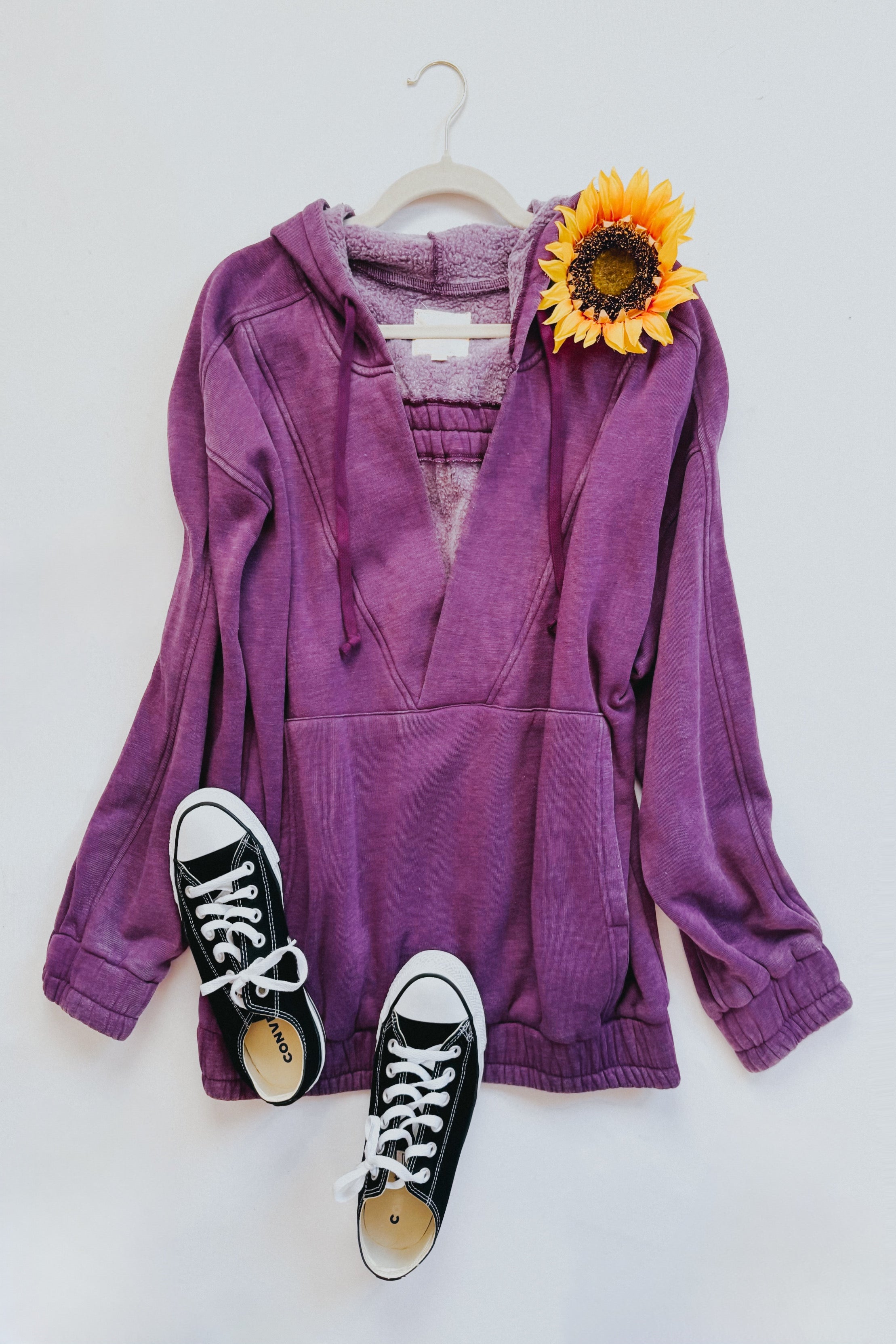 Plum acid wash hoodie with plush lining hanging with black sneakers and sunflower accent on white wall background