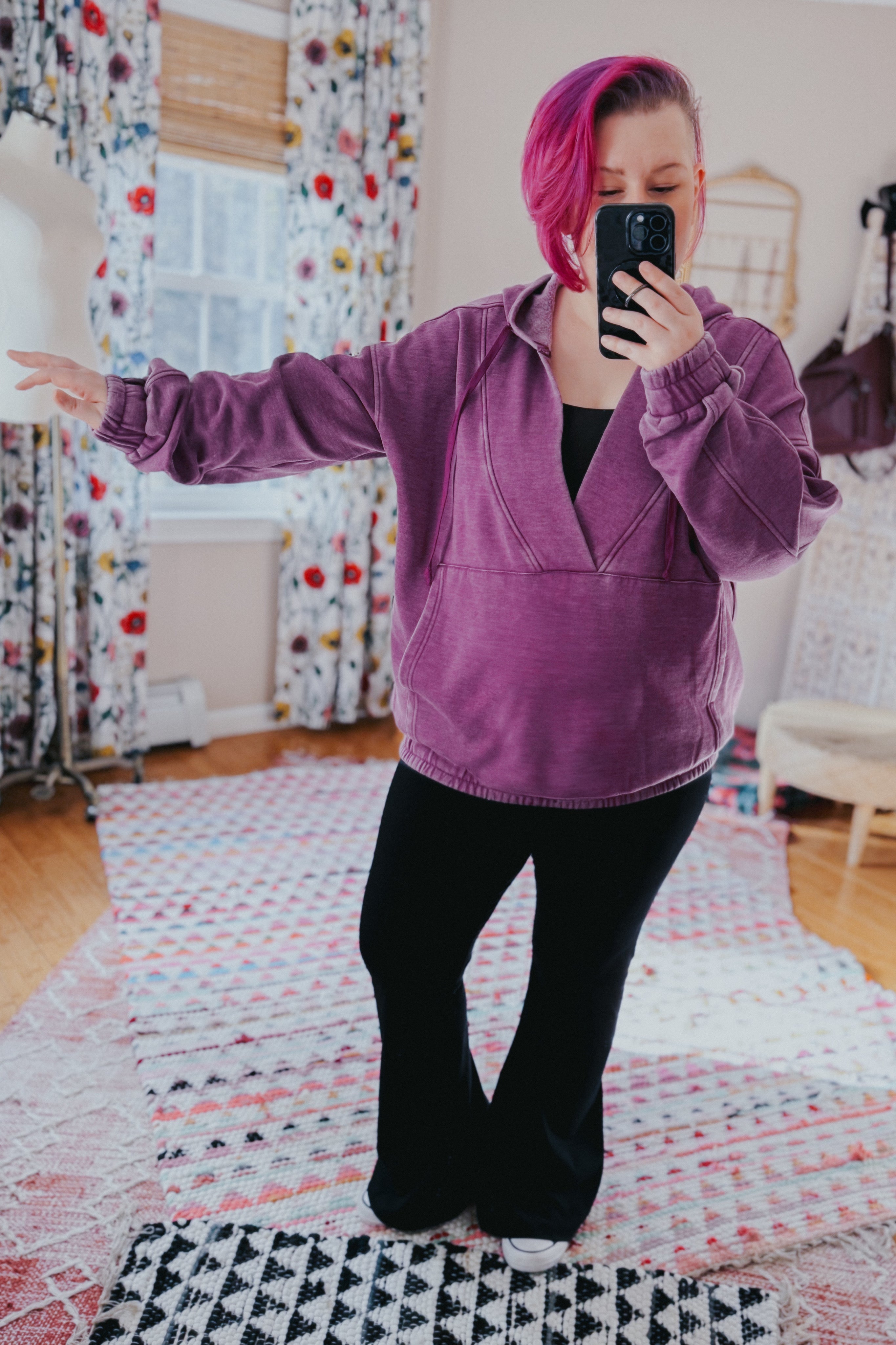 Woman wearing plum acid wash hoodie with gathered cuffs taking a mirror selfie in cozy, colorful room setting