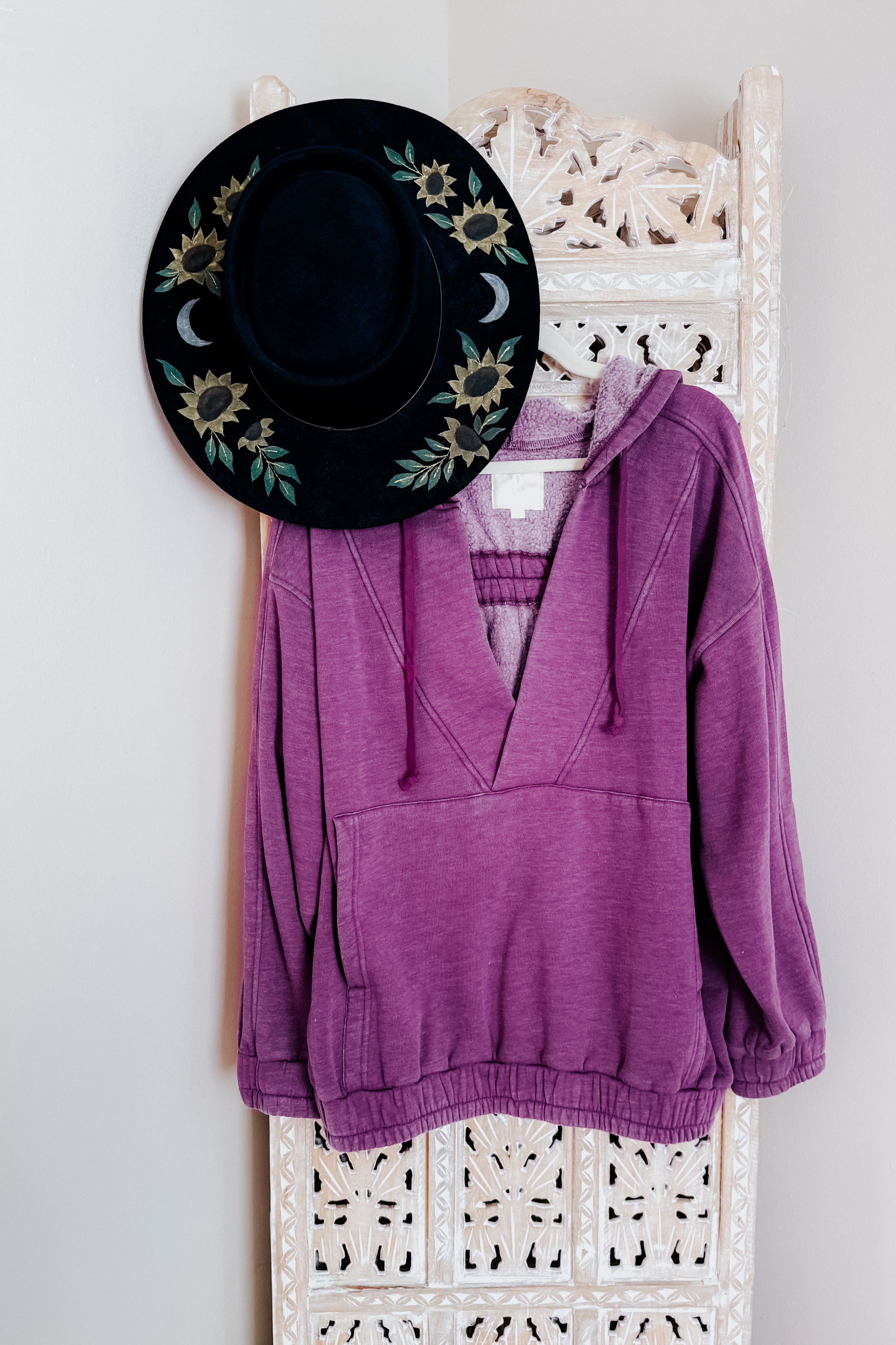 Plum acid wash hoodie displayed on carved wooden panel with black hat featuring floral and moon designs