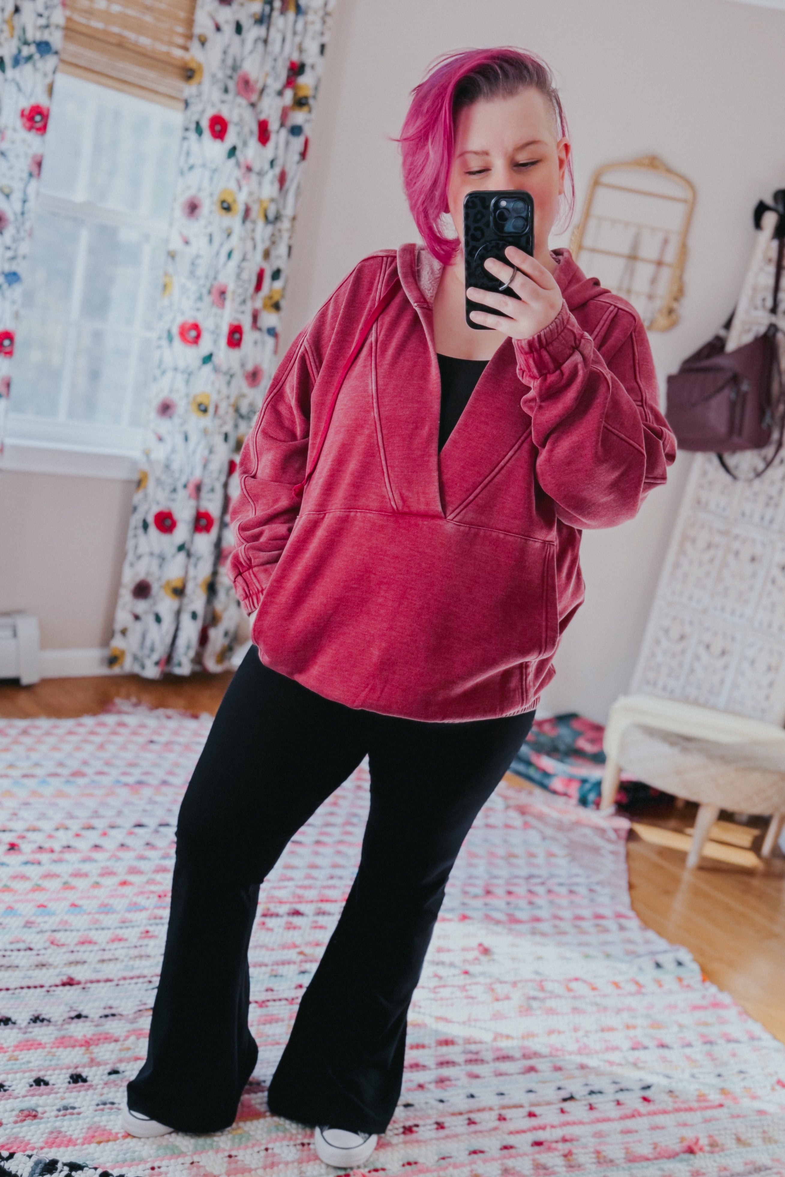 Woman with short magenta hair wearing wine acid wash hoodie and black flared pants taking an indoor mirror selfie