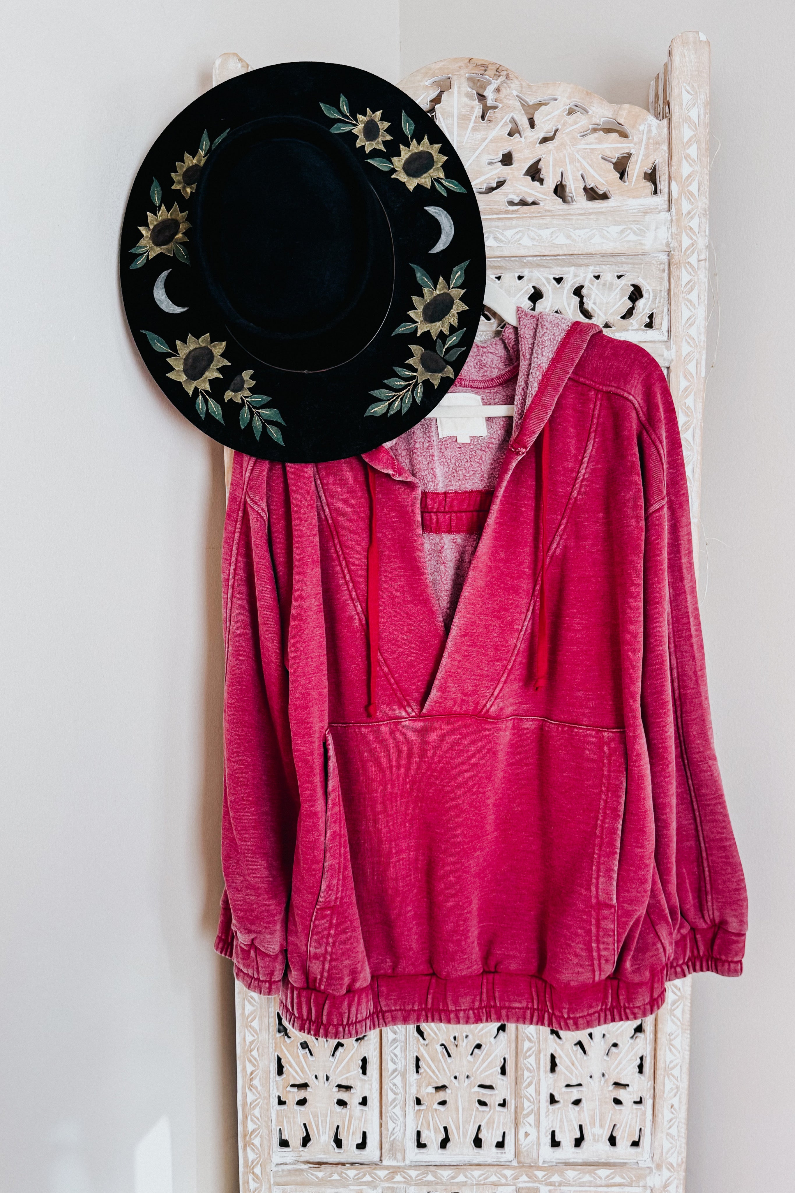 Wine-colored acid wash hoodie with hood and front kangaroo pocket hanging on carved white wooden screen with black hat adorned with floral and moon embroidery