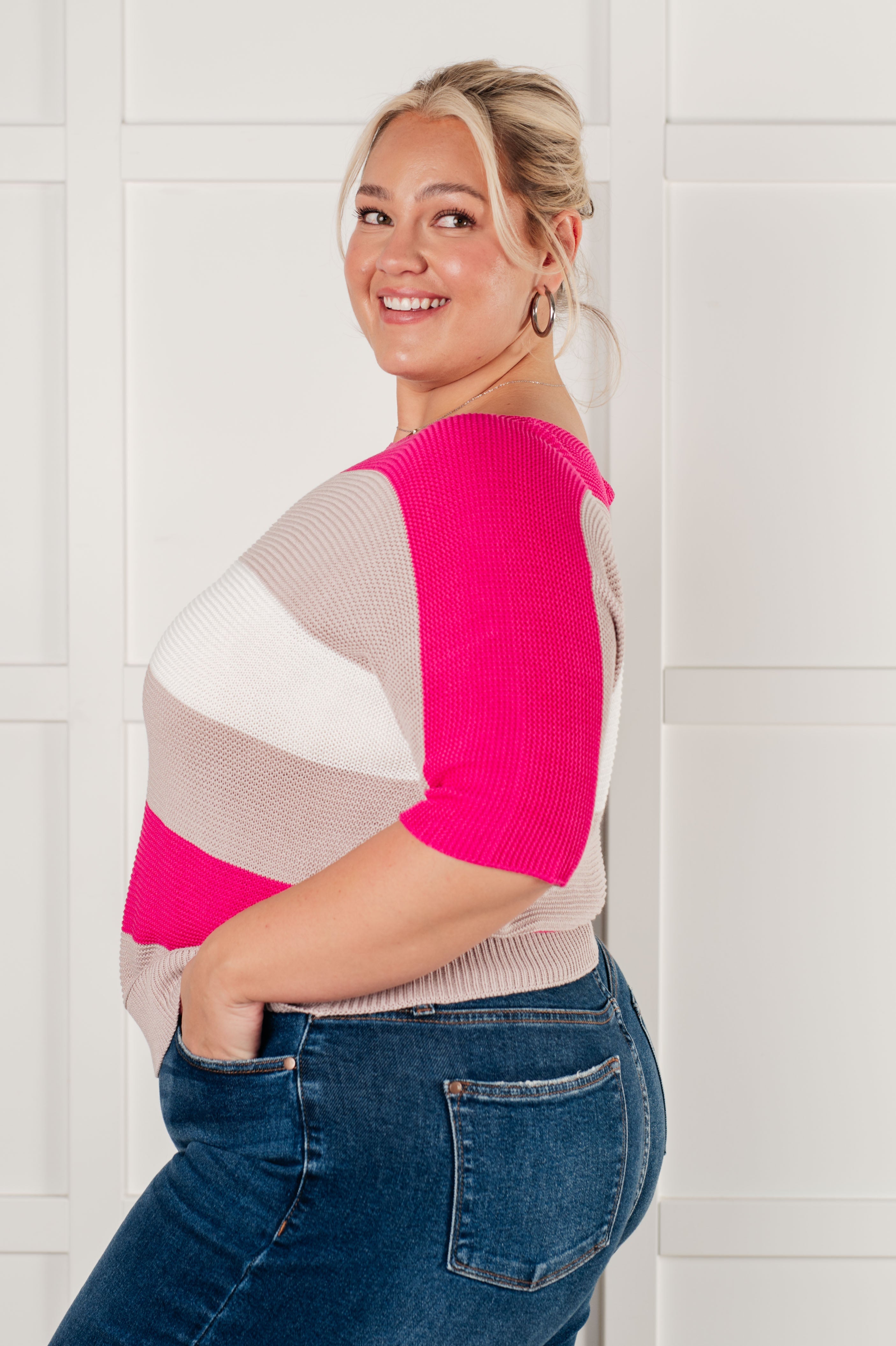 Smiling woman wearing color-blocked pink and beige short sleeve knit top with high-waisted blue jeans standing sideways