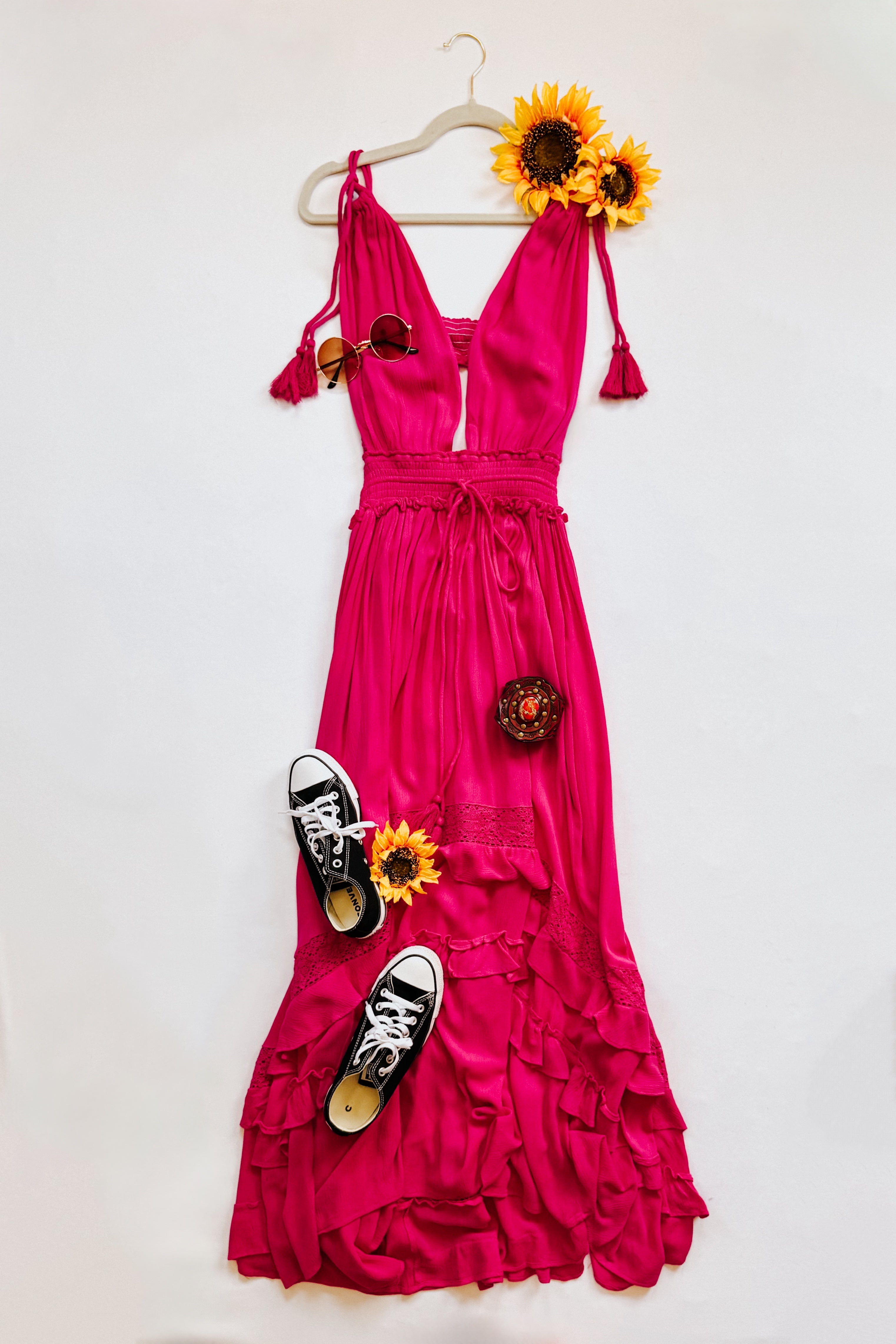 Fuchsia maxi dress with ruffle details styled with black sneakers, sunglasses, and sunflower accents on a hanger