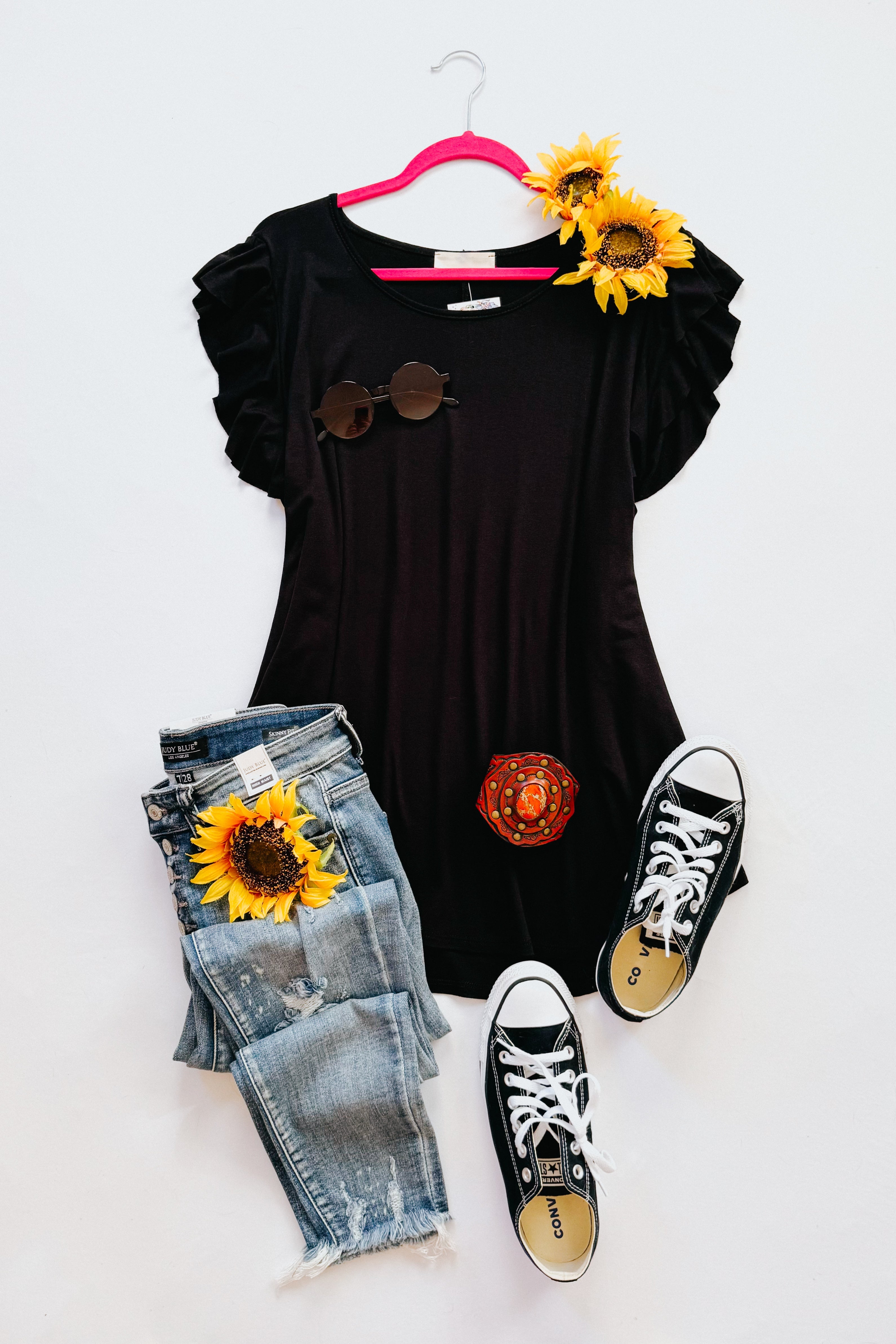 Black ruffle sleeve top paired with distressed denim jeans, black sneakers, sunglasses, and sunflower accents on white background