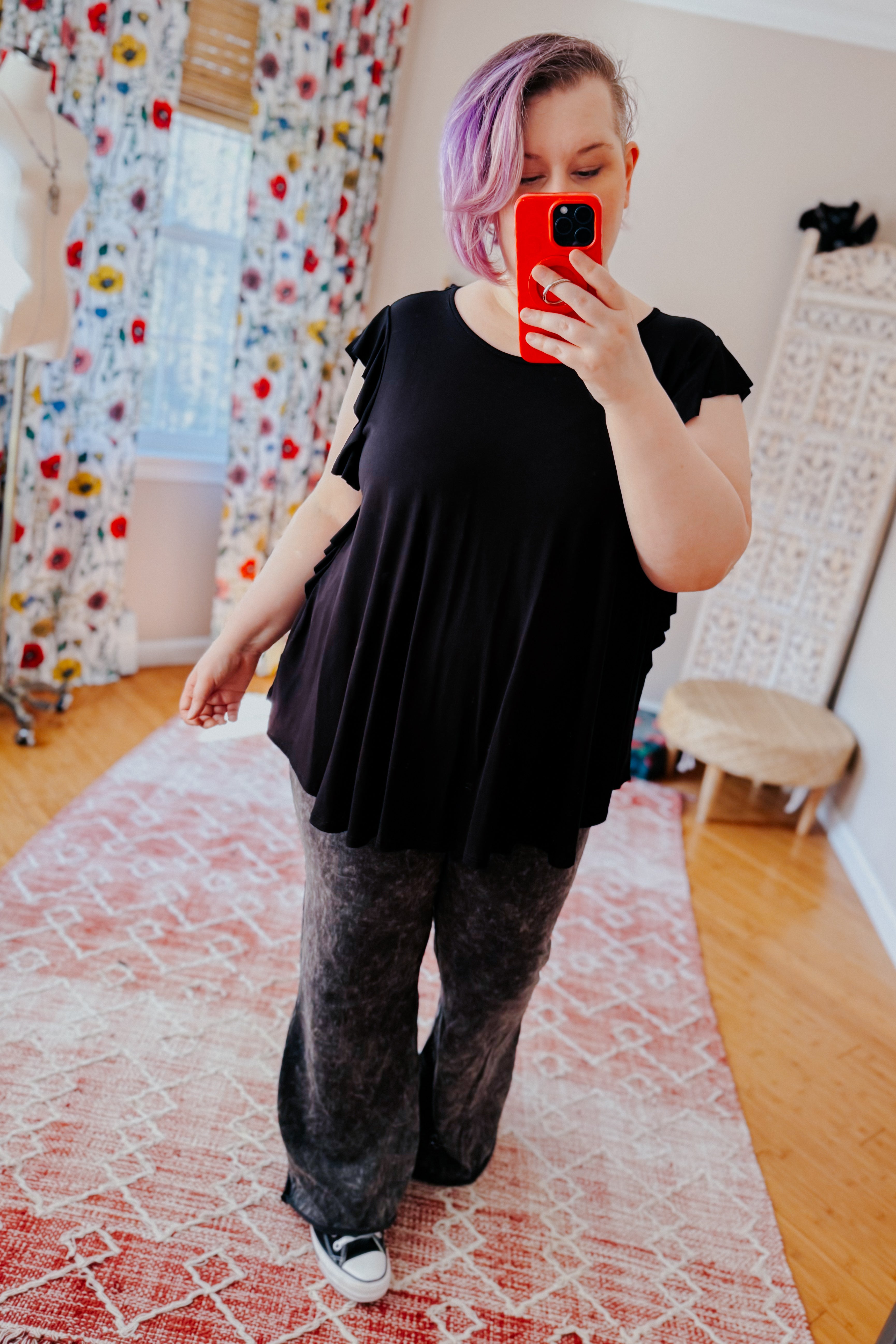 Woman wearing black ruffle sleeve top and wide-leg pants taking selfie in bright room with colorful floral curtains