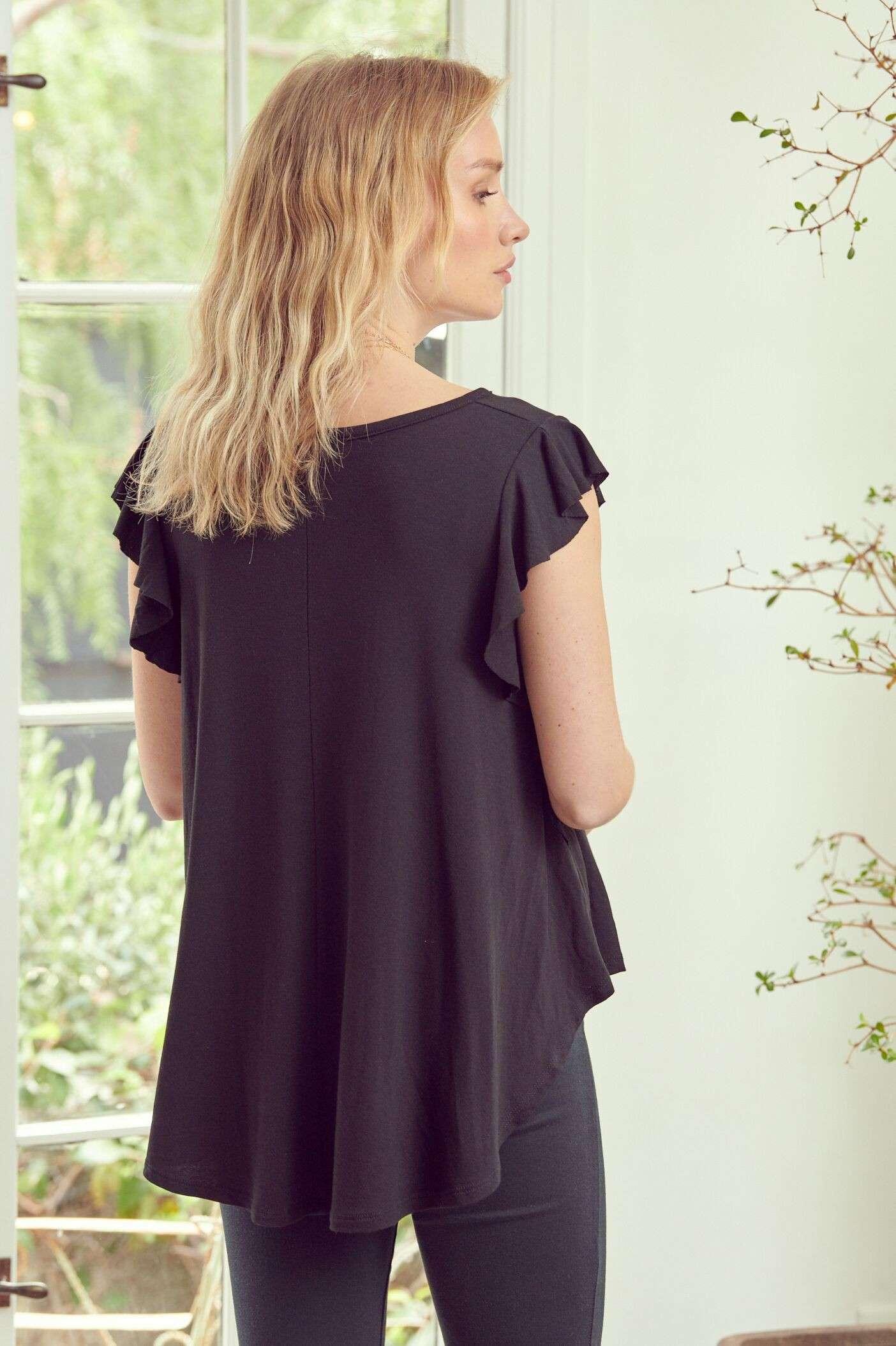 Woman wearing black ruffle sleeve top with loose fit standing indoors near window with natural light and plants