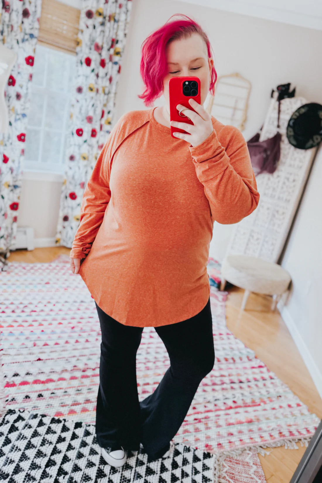 Woman with pink hair wearing orange rustic charm long sleeve top and black flare pants taking mirror selfie indoors