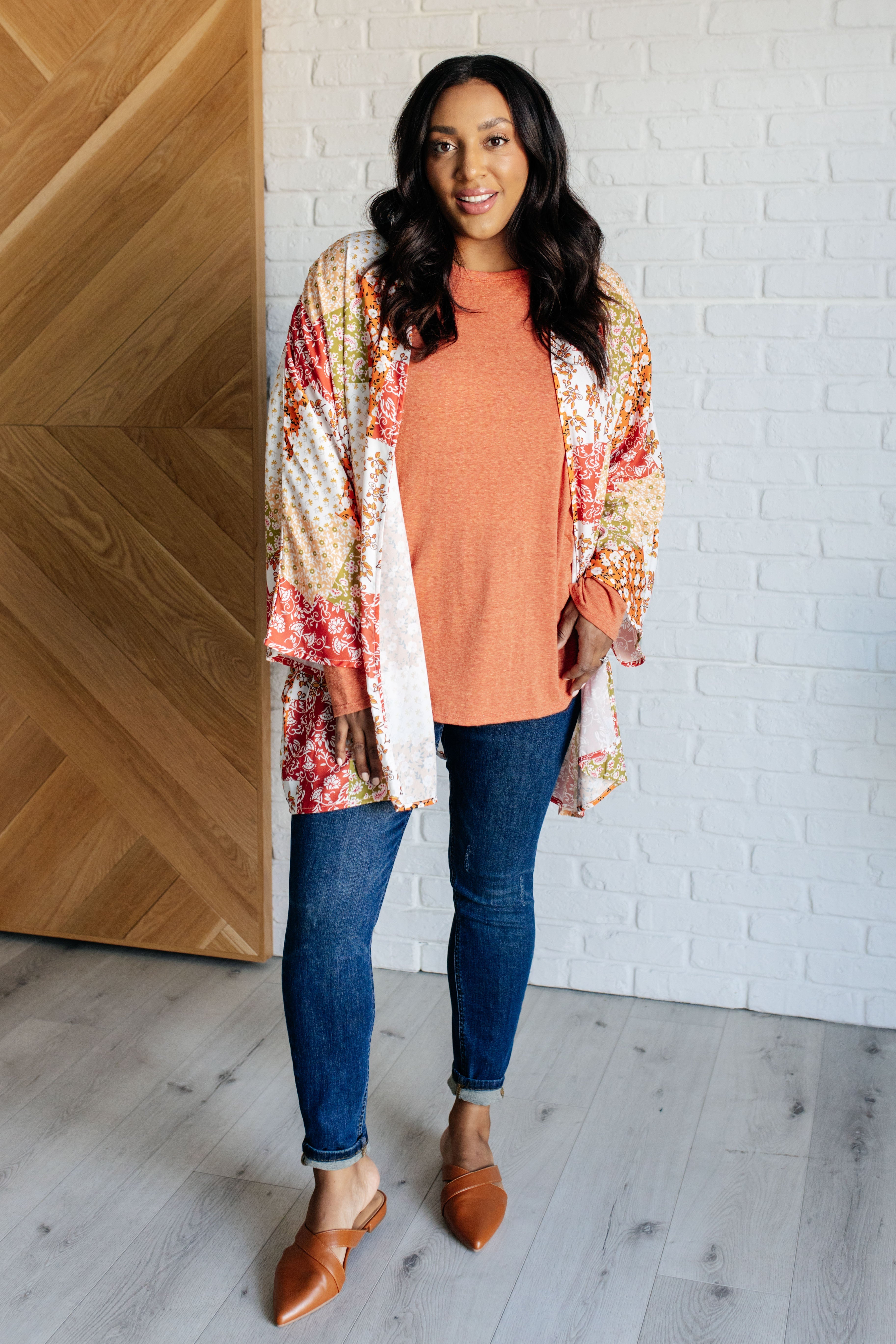 Woman wearing orange long sleeve top under colorful patchwork kimono paired with slim dark jeans and brown flats indoors