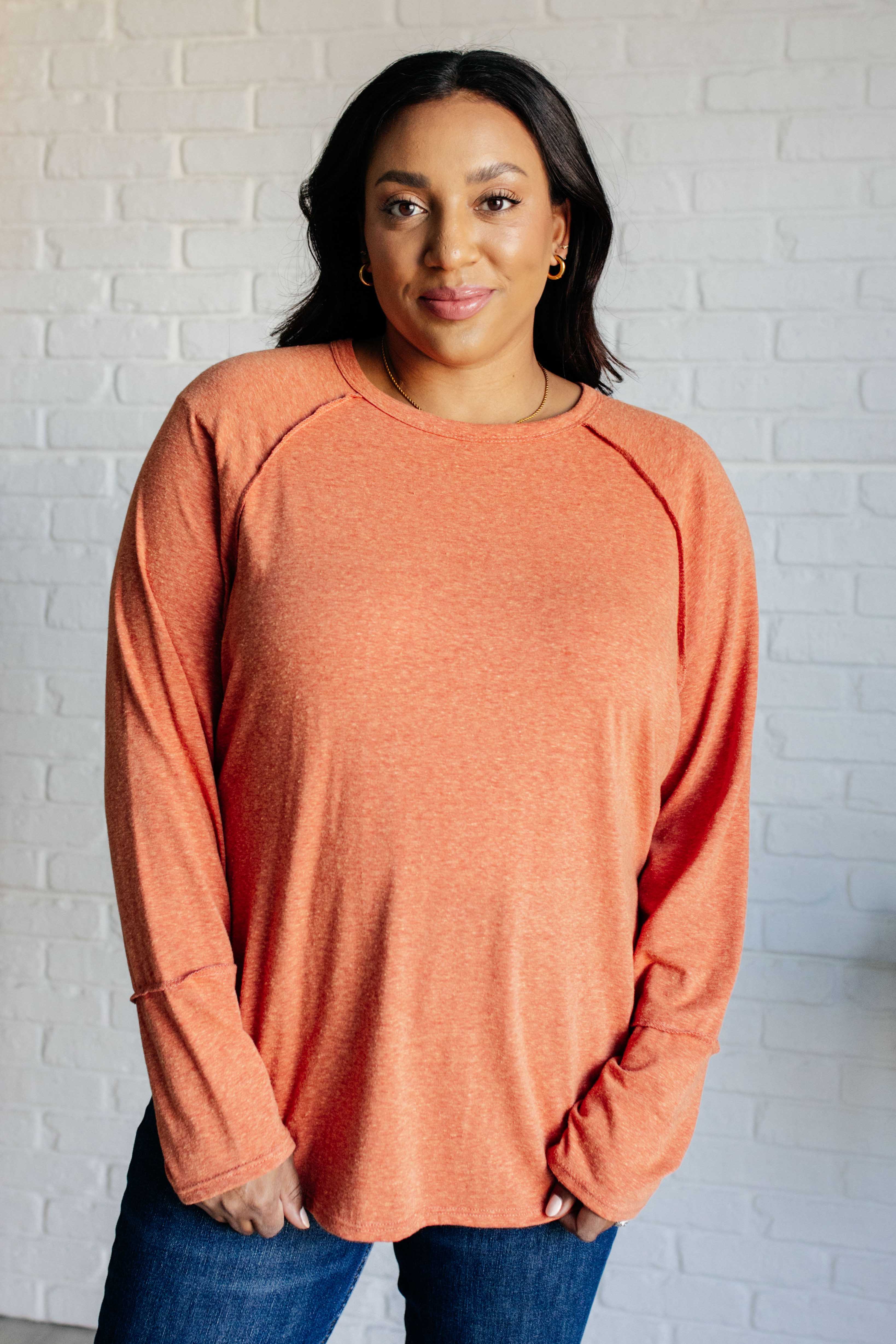 Woman wearing orange long sleeve top with relaxed fit and crew neckline standing against white brick wall