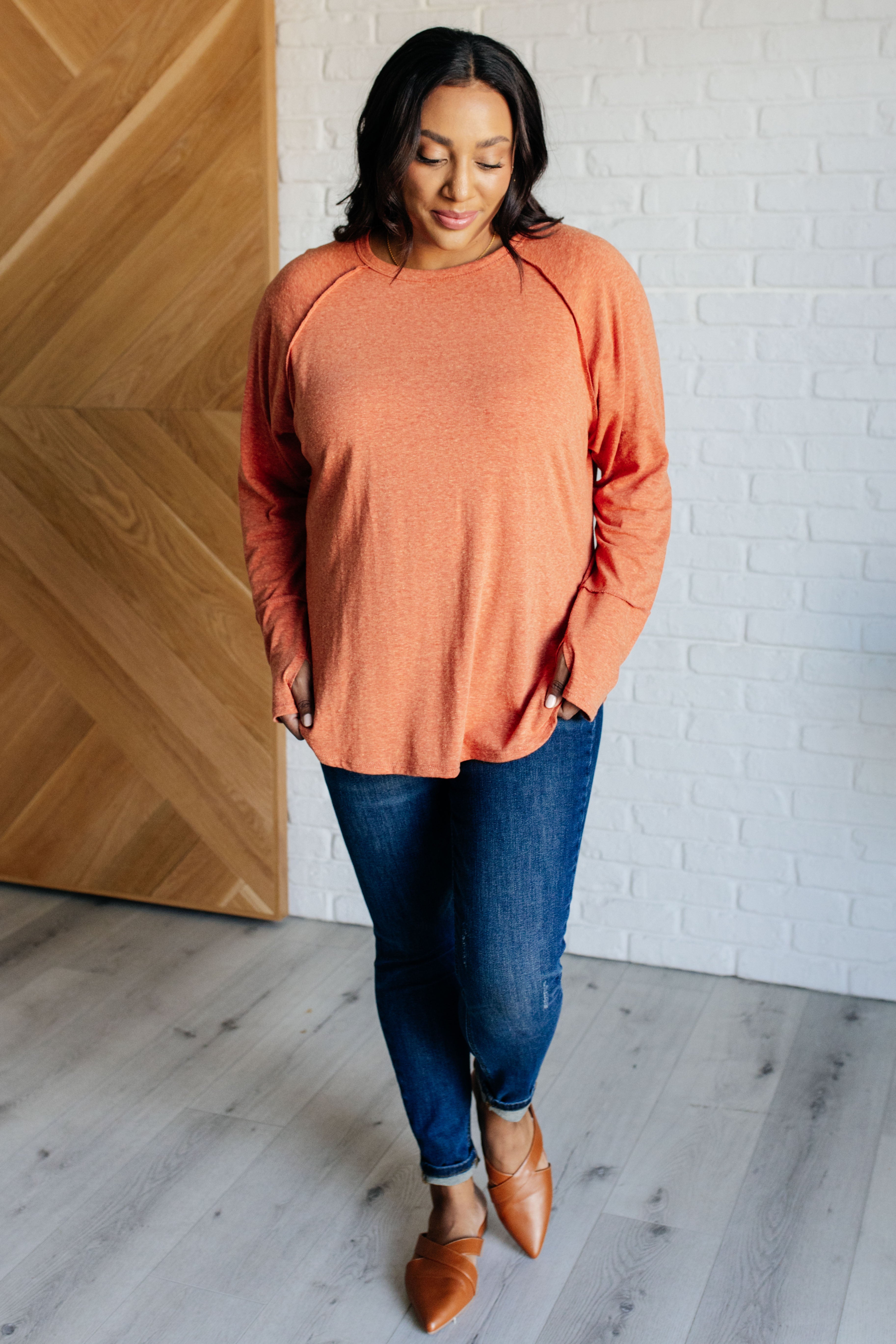 Woman wearing orange long sleeve top with relaxed fit paired with slim dark blue jeans and brown pointed flats indoors