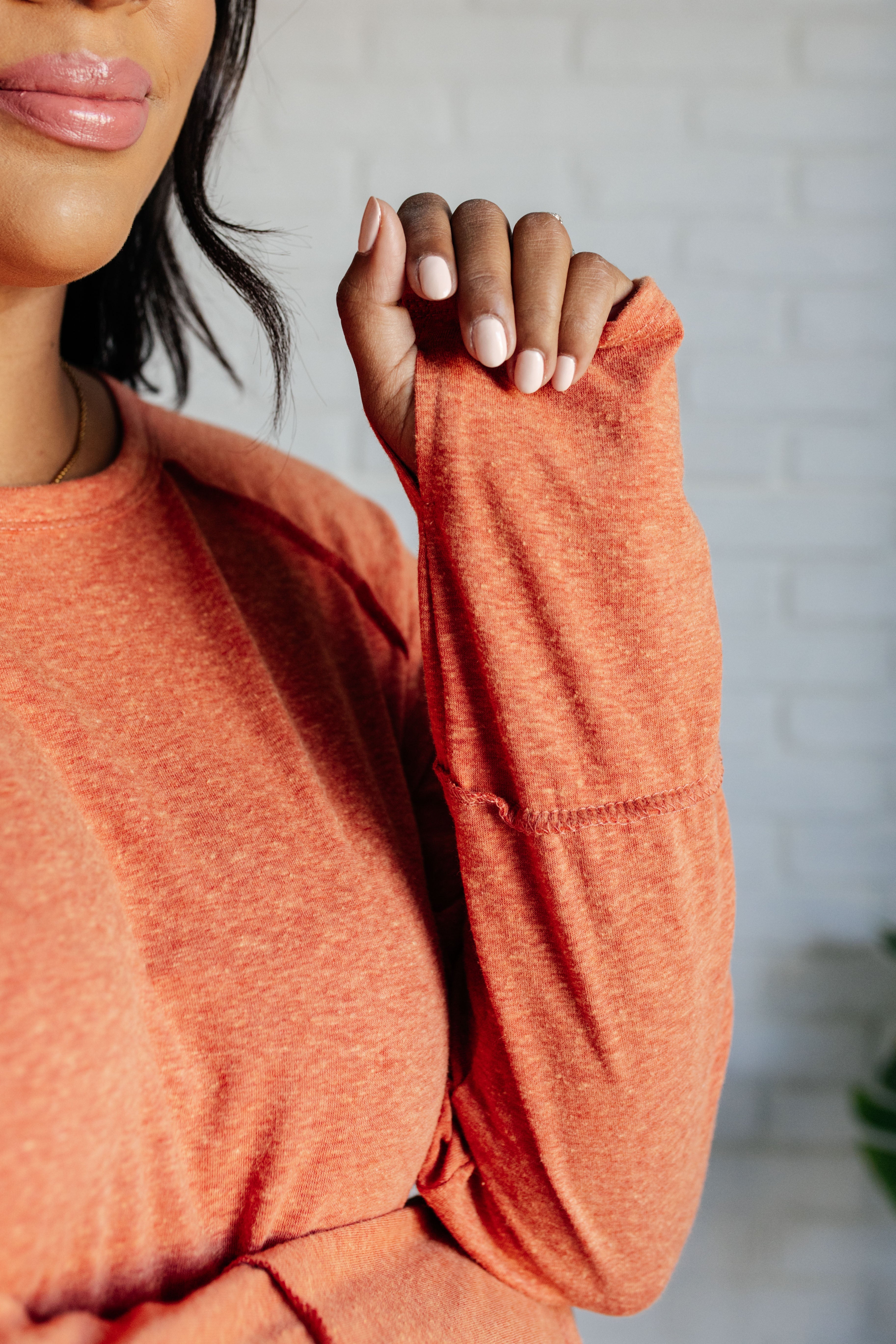 Close-up of person wearing soft orange long sleeve top with thumb hole detail against light brick wall background