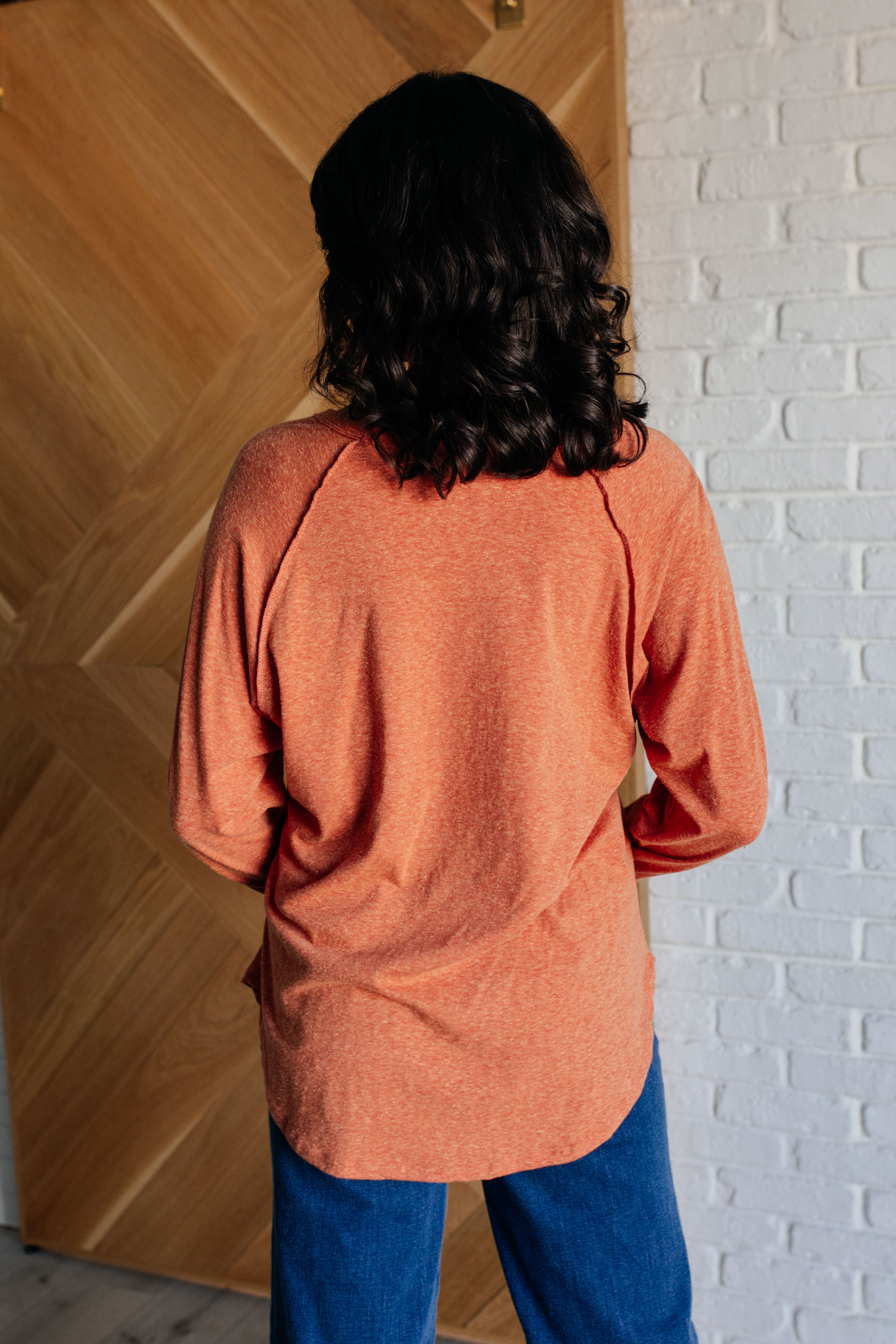 Person wearing relaxed fit orange long sleeve top standing with back facing camera indoors near wooden and white brick walls