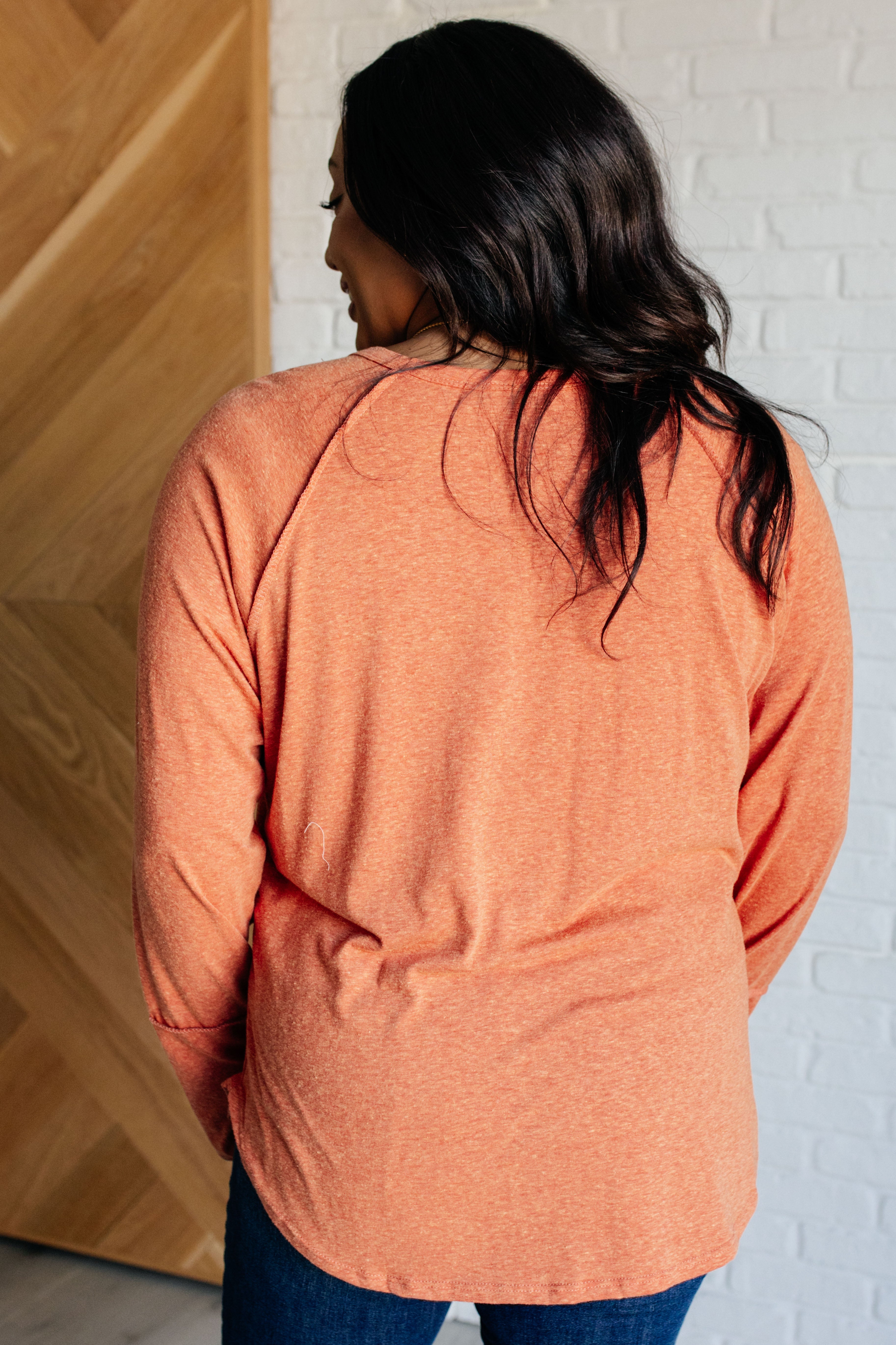 Woman wearing soft orange long sleeve top with relaxed fit standing indoors against wood and white brick wall