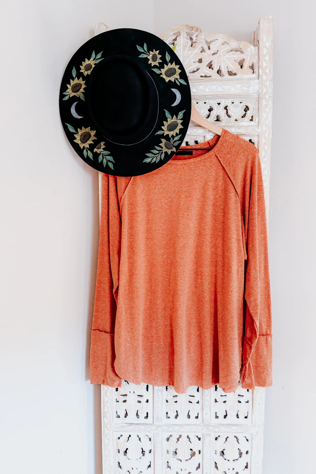 Orange long sleeve top with relaxed fit hanging on carved white wooden panel alongside black wide-brim hat with sunflower and moon details