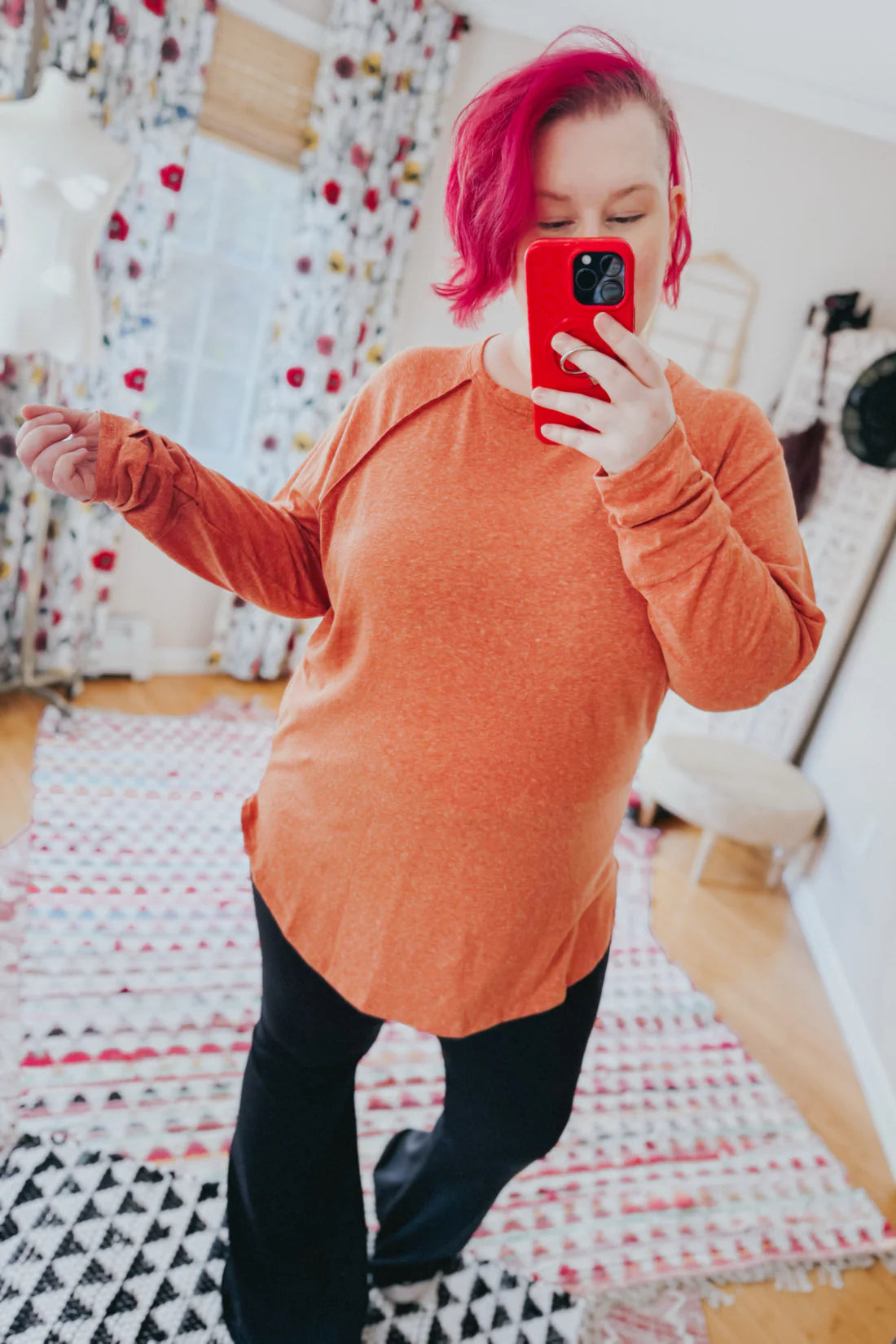 Woman with pink hair wearing orange rustic charm long sleeve top and black pants taking indoor selfie
