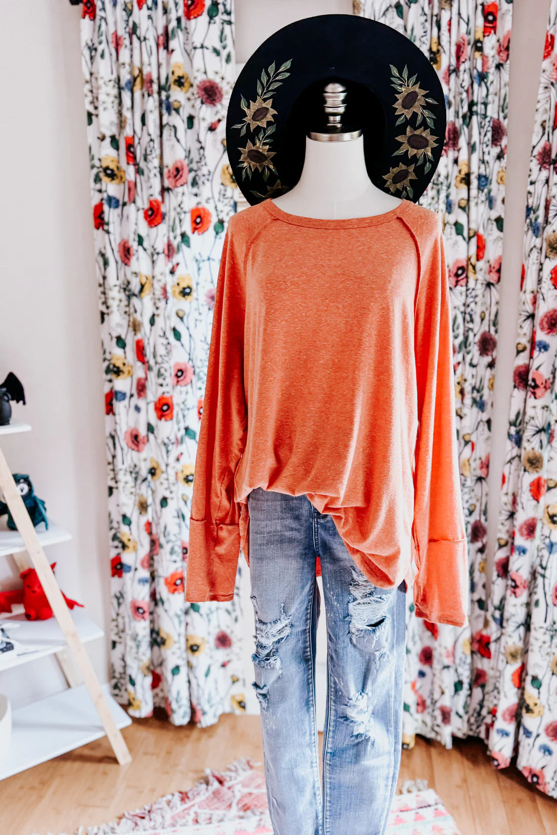 Orange long sleeve top paired with distressed jeans on mannequin with floral curtains and black hat background