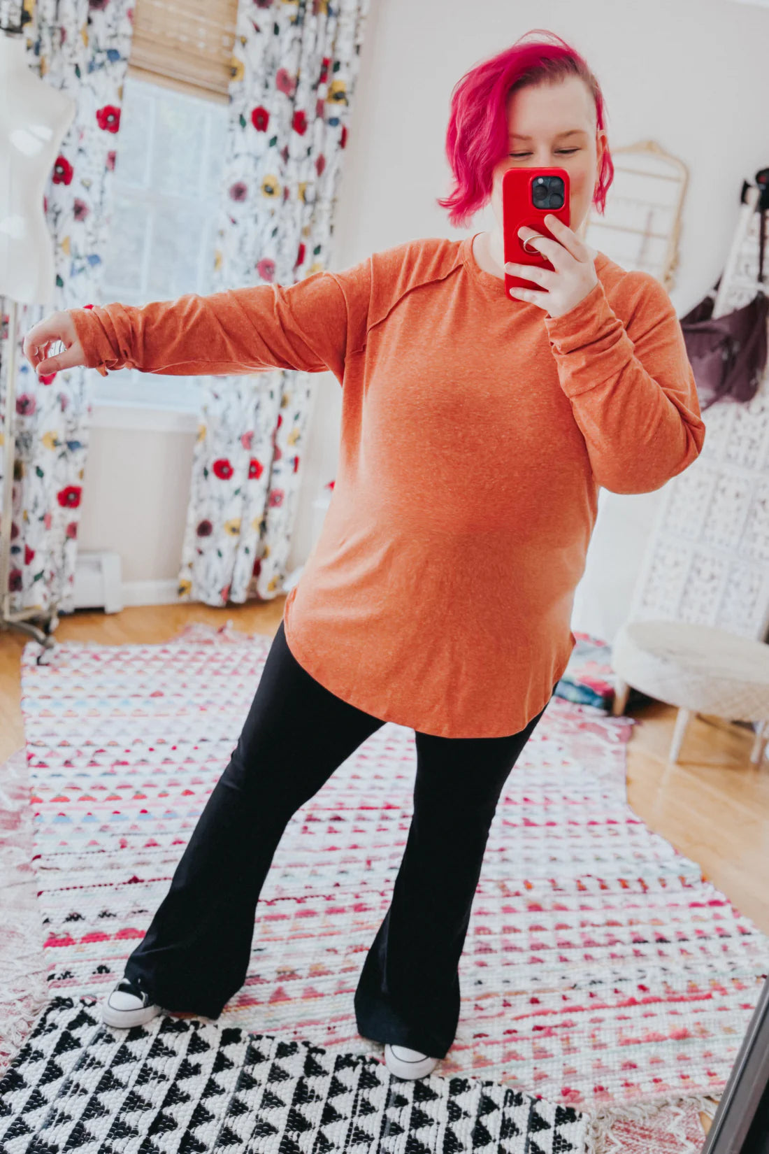 Woman with pink hair wearing orange long sleeve top and black flared pants taking mirror selfie indoors