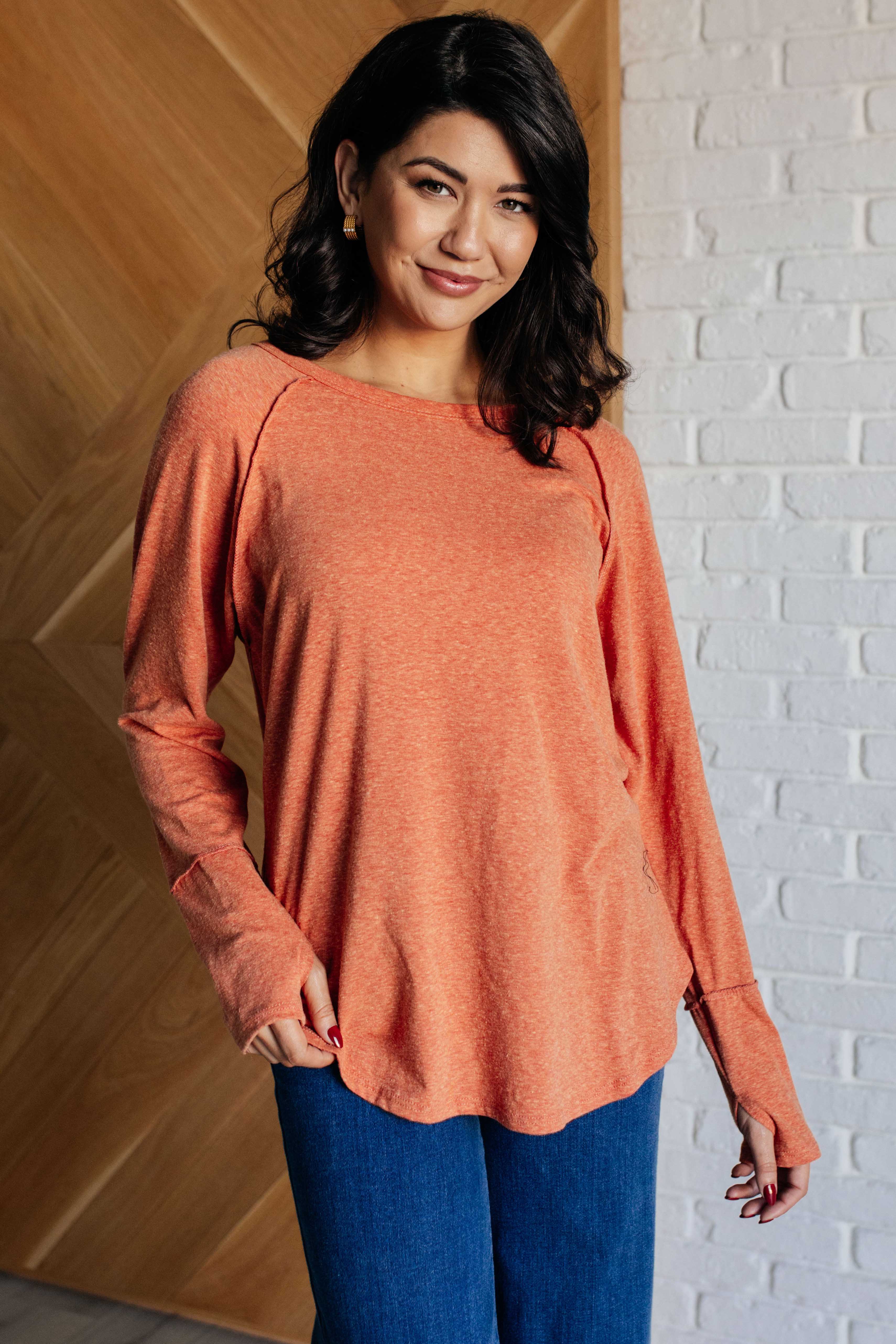 Woman wearing rustic orange long sleeve top with thumb holes paired with blue jeans indoors against wood and brick wall