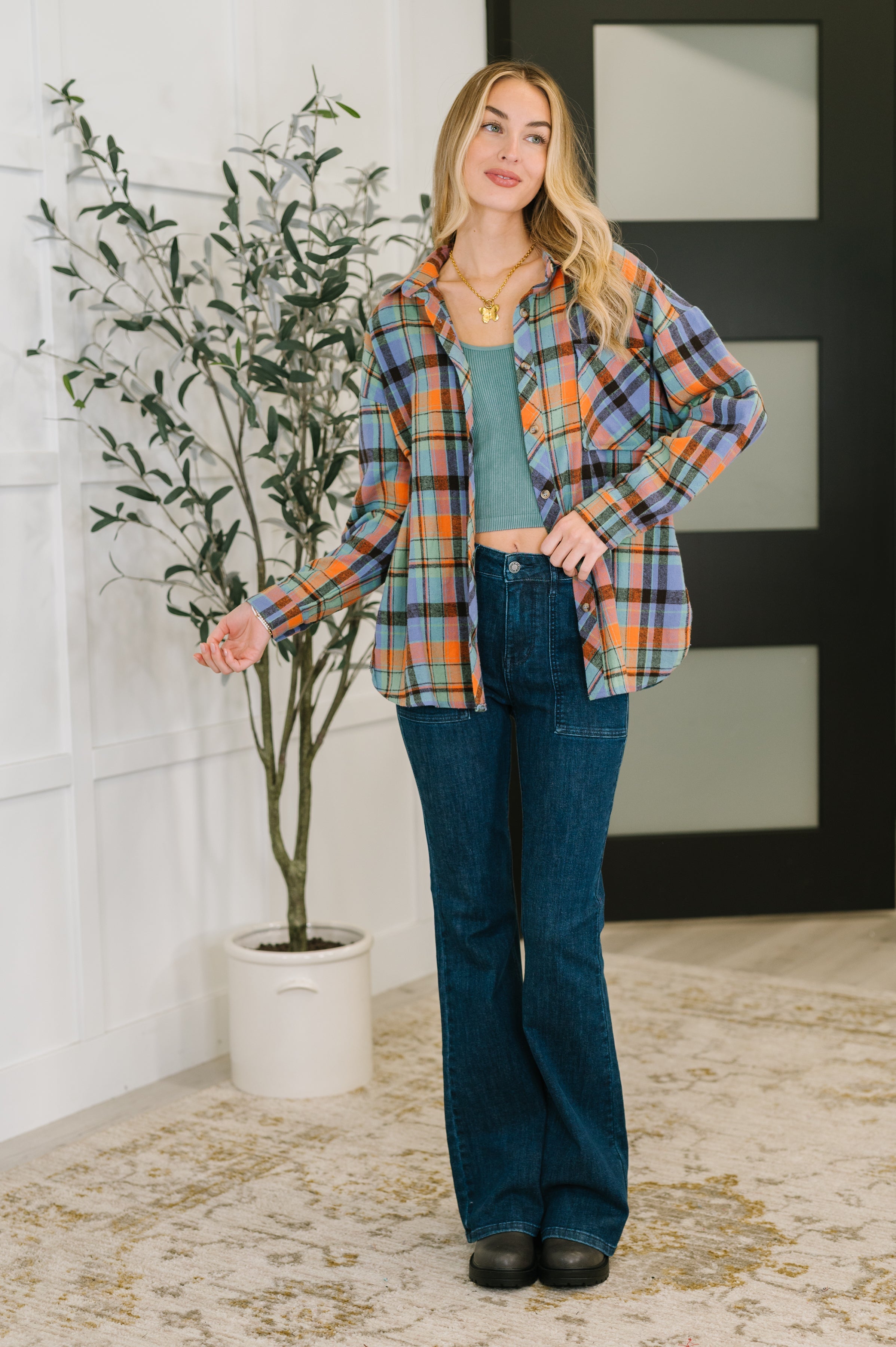 Woman wearing multicolored plaid button down over blue tank top paired with dark blue flared jeans and black shoes indoors