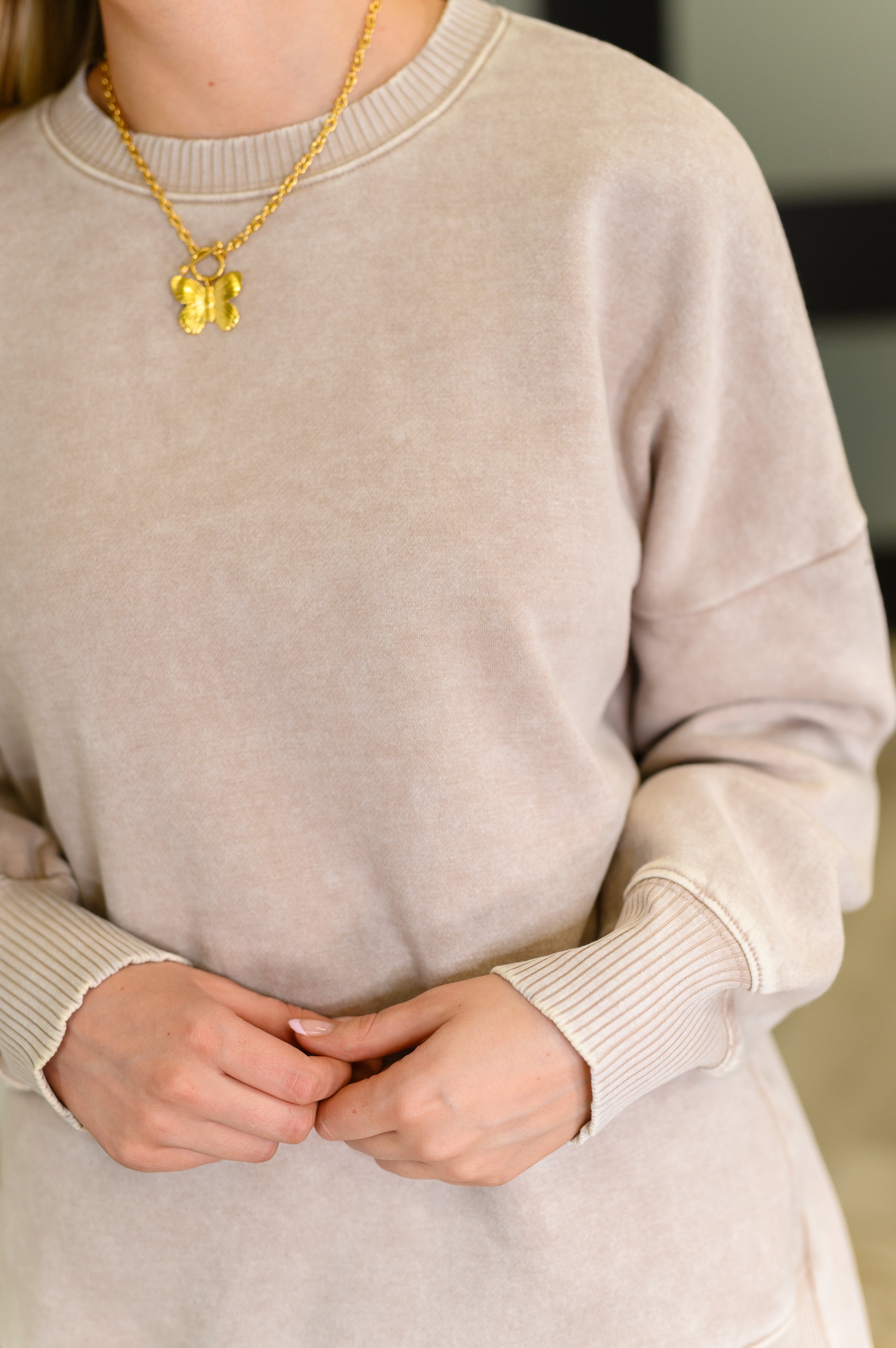 Close-up of woman wearing ash mocha mineral wash pullover with ribbed cuffs and gold butterfly necklace
