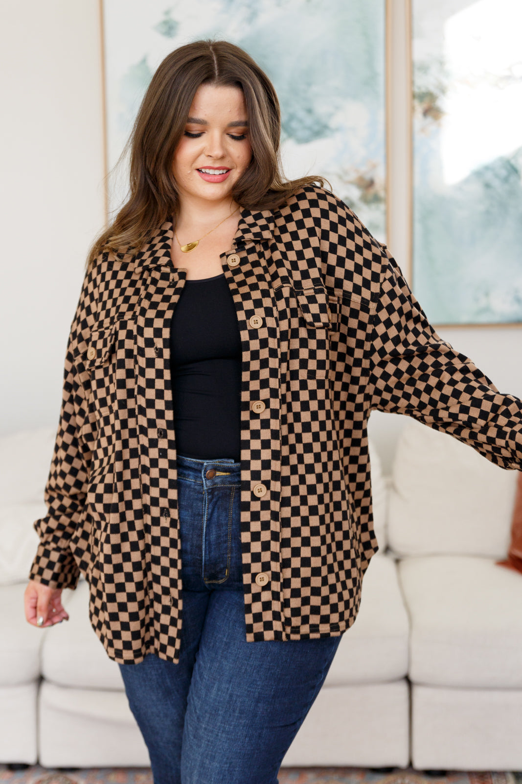 Woman wearing brown checkered button-up shacket over black top and jeans standing indoors near white couch