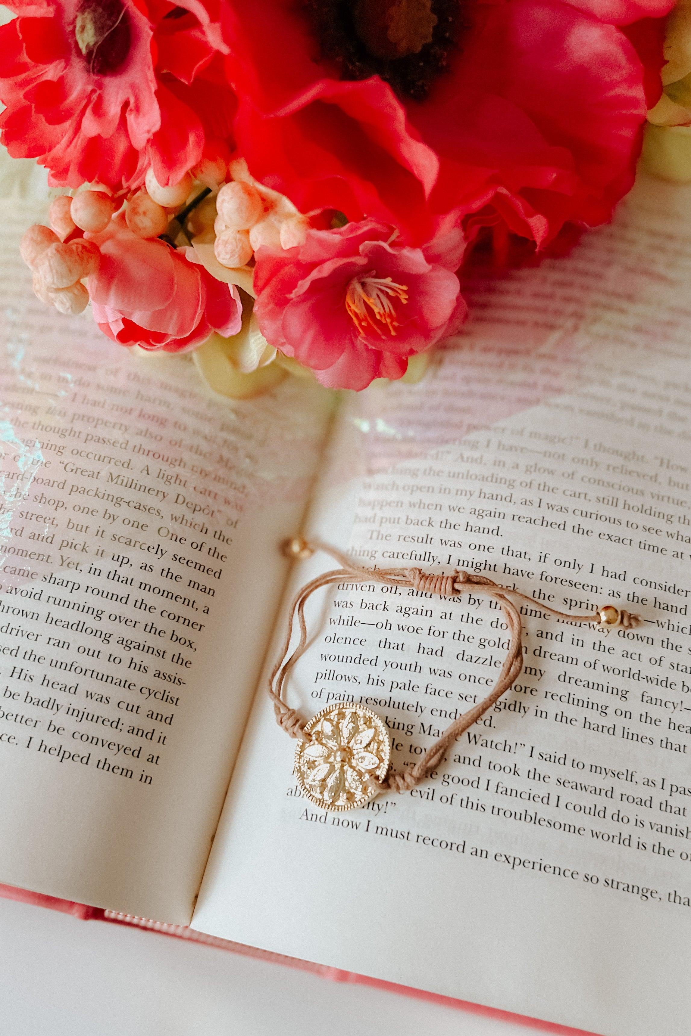 Beige stackable bracelets with intricate charms resting on an open book beside vibrant red flowers.
