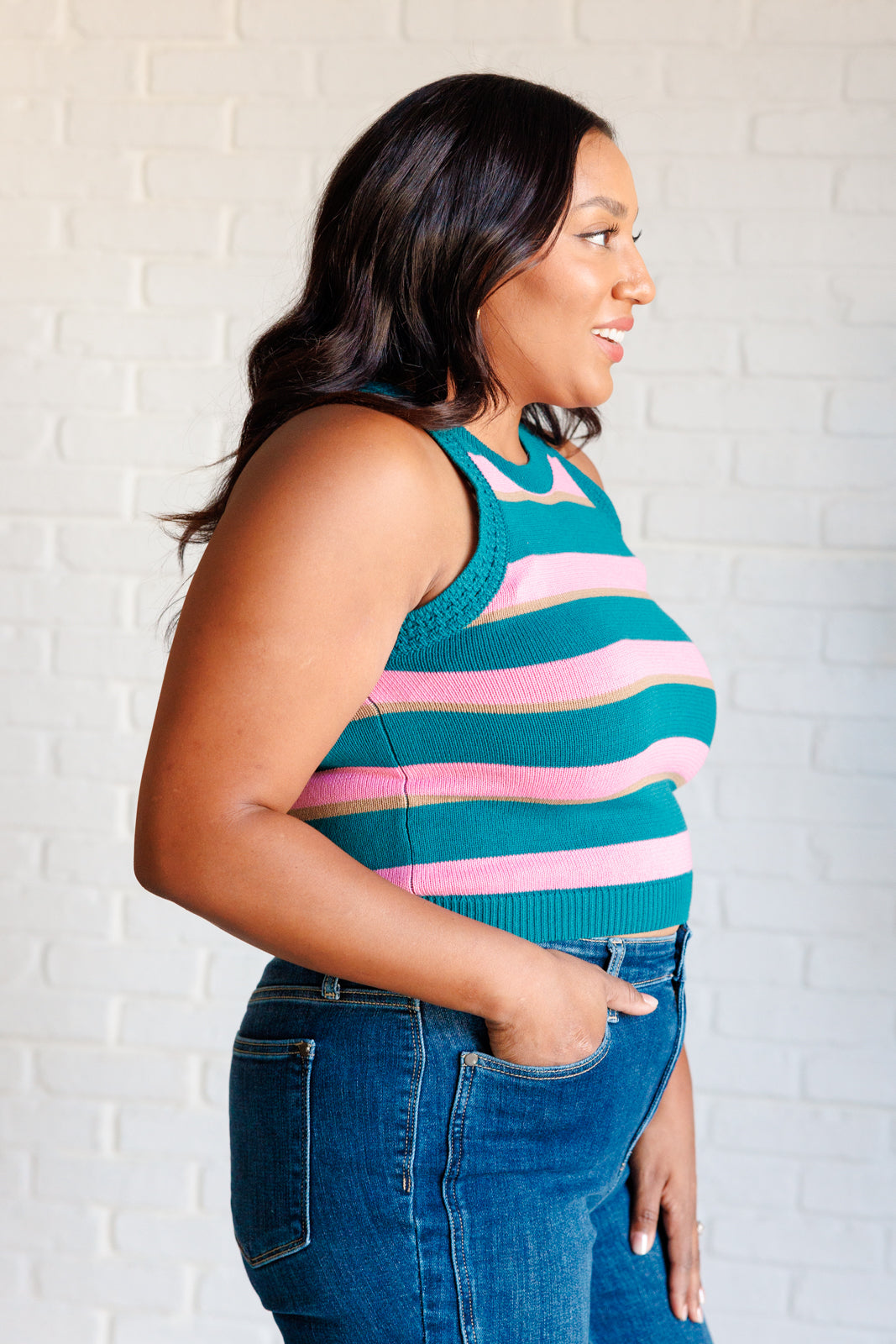 Side profile of woman wearing blue knit tank with pink and green stripes paired with medium wash jeans indoors