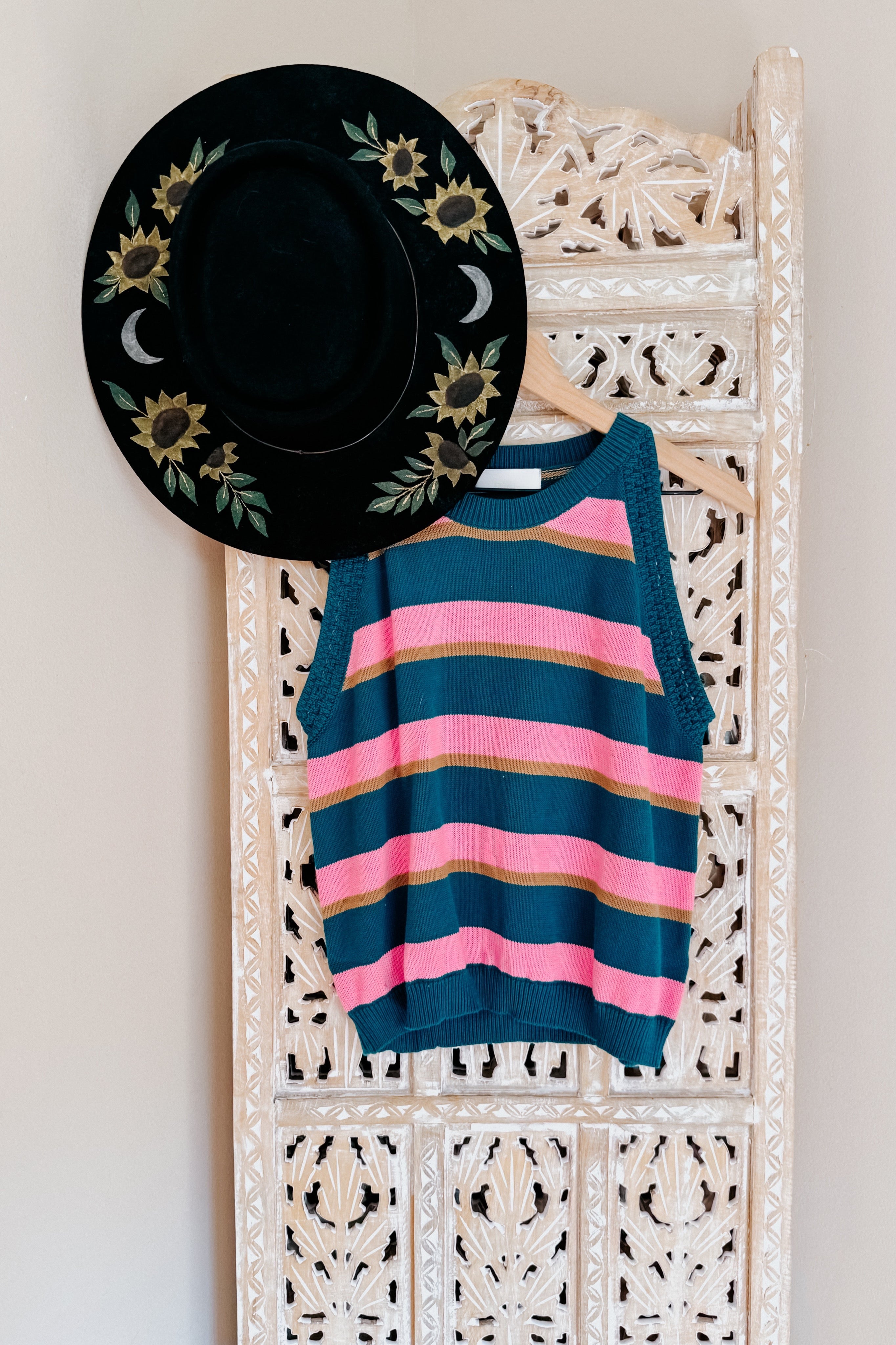 Blue knit striped tank with pink and tan stripes hanging on carved wooden panel alongside black hat with floral and moon embroidery