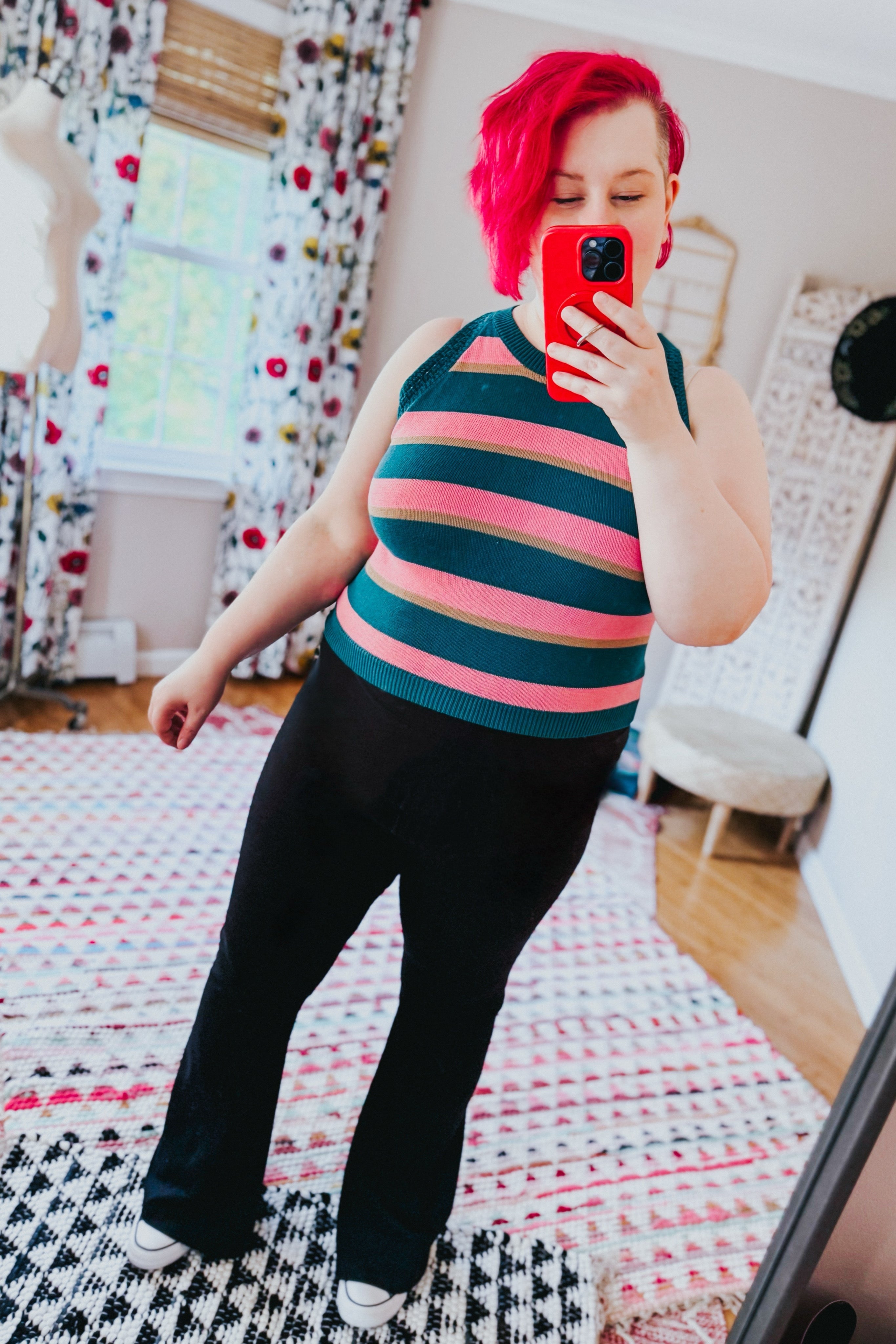 Woman with red hair wearing a striped sleeveless knit top paired with black pants taking a mirror selfie indoors
