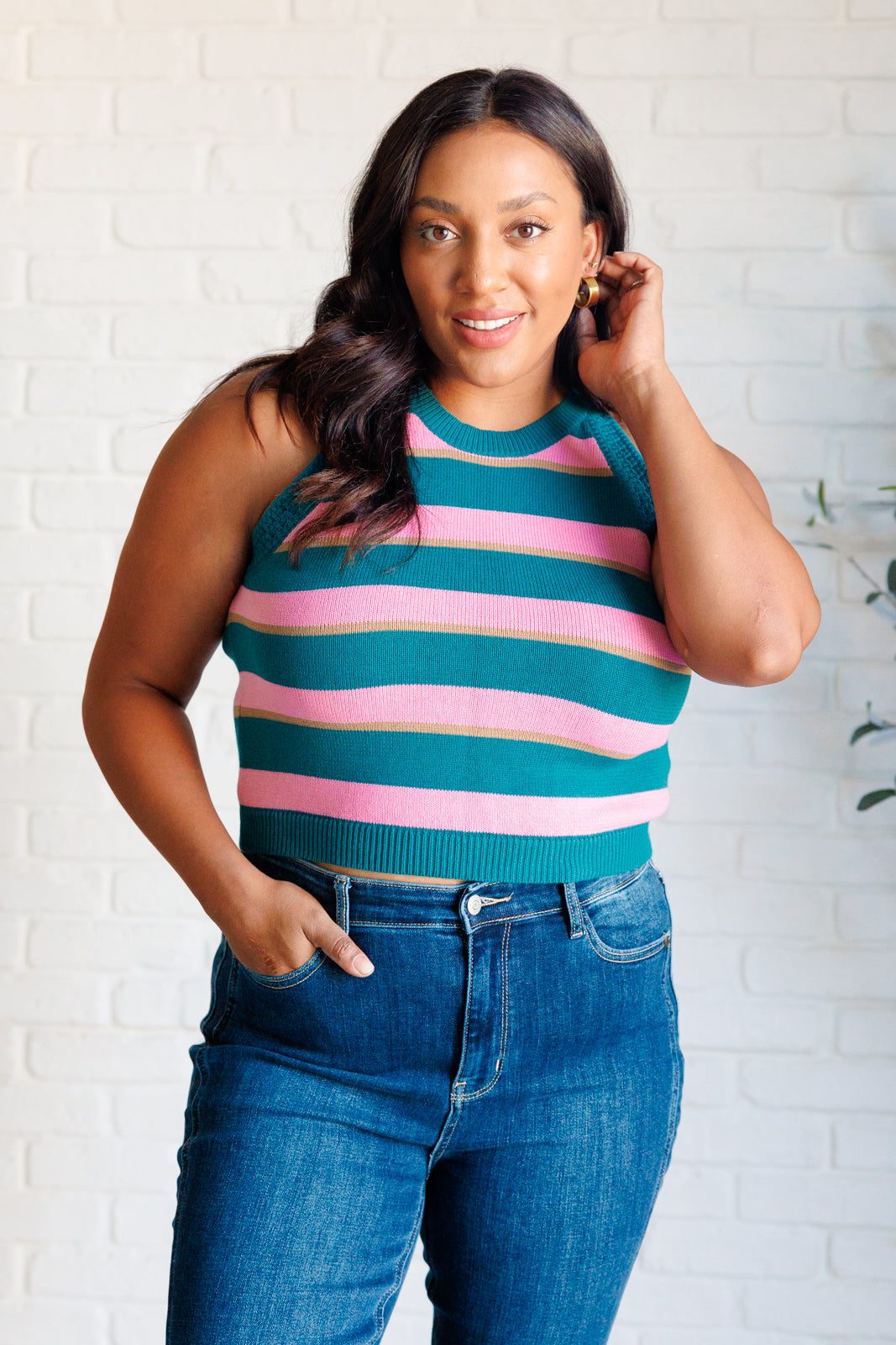 Woman wearing blue and pink striped knit tank paired with high-waisted blue jeans standing against white brick wall