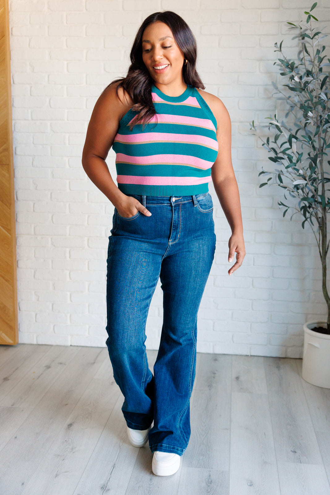 Woman wearing green and pink striped knit tank paired with high-waisted blue jeans and white sneakers indoors