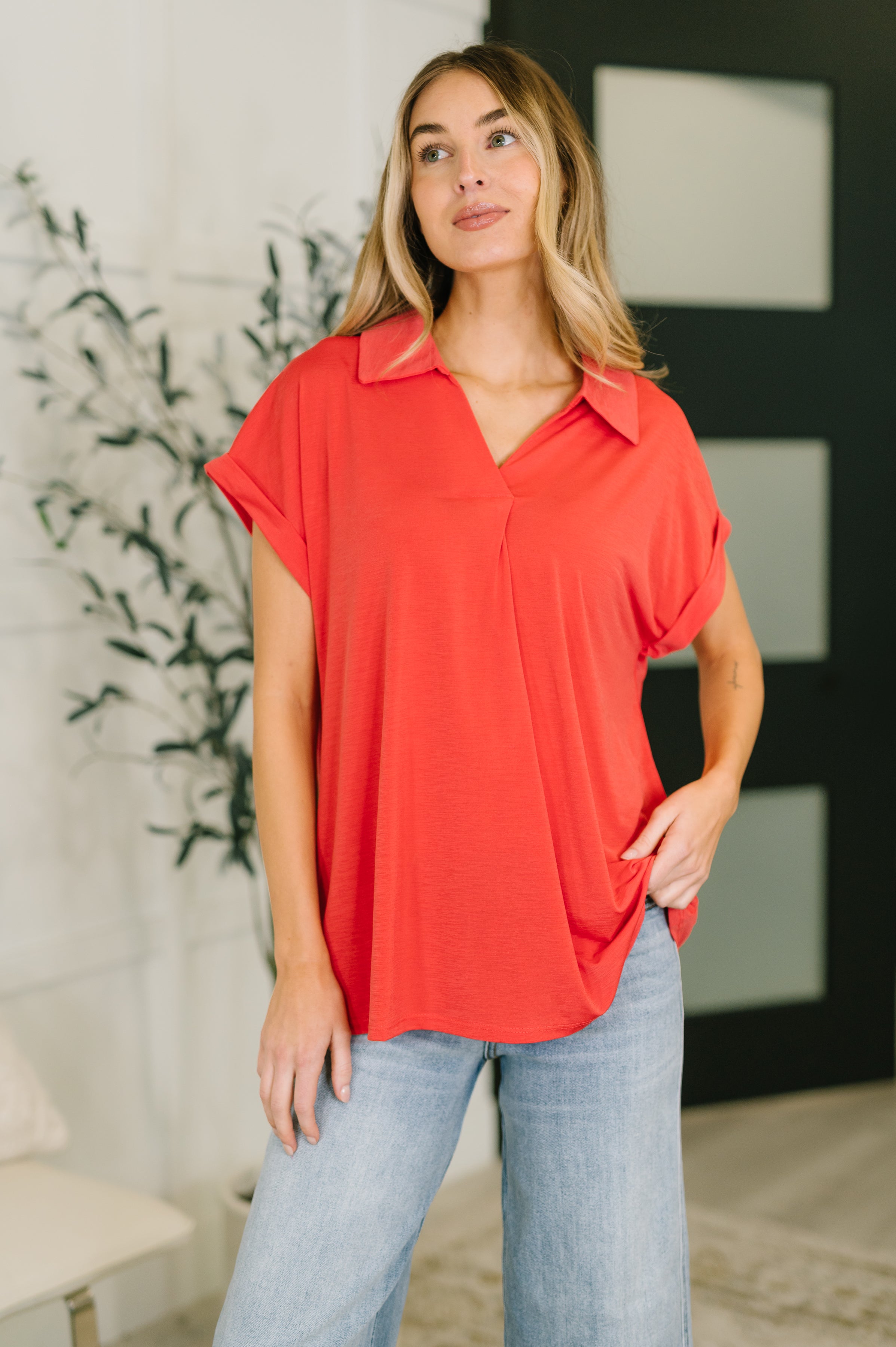 Woman wearing loose hot coral collared top with dolman sleeves and light wash jeans indoors near plant and black door panels