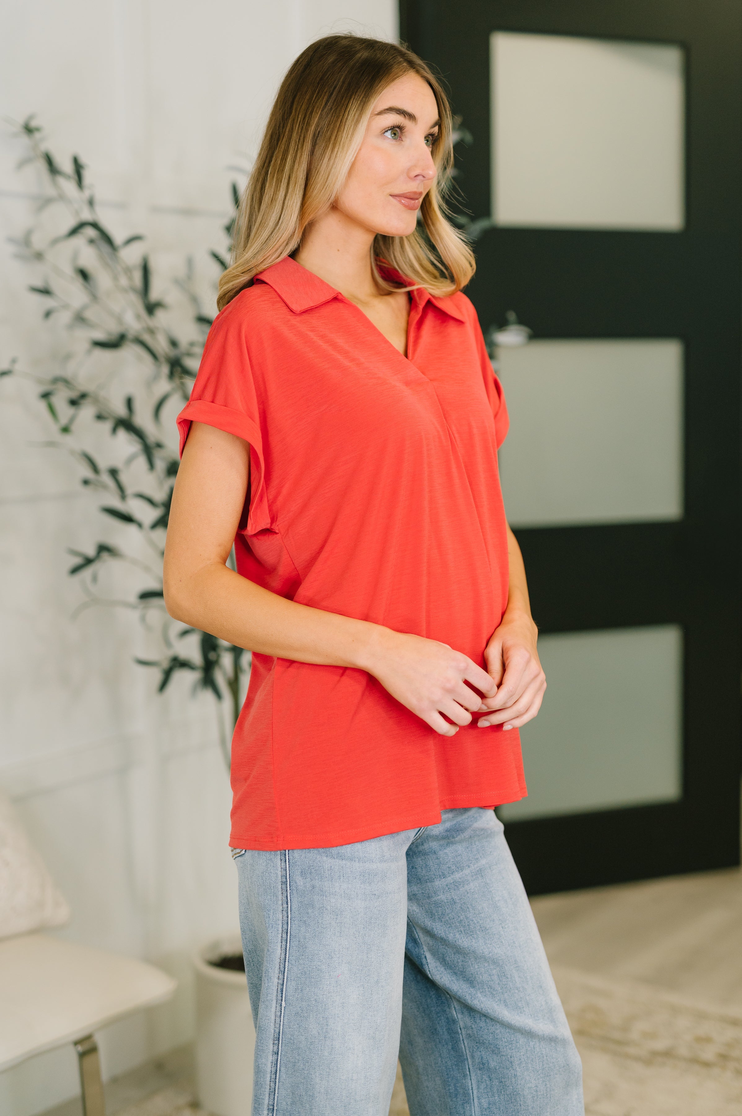 Woman wearing relaxed coral collared top with cuffed short sleeves paired with light blue jeans indoors