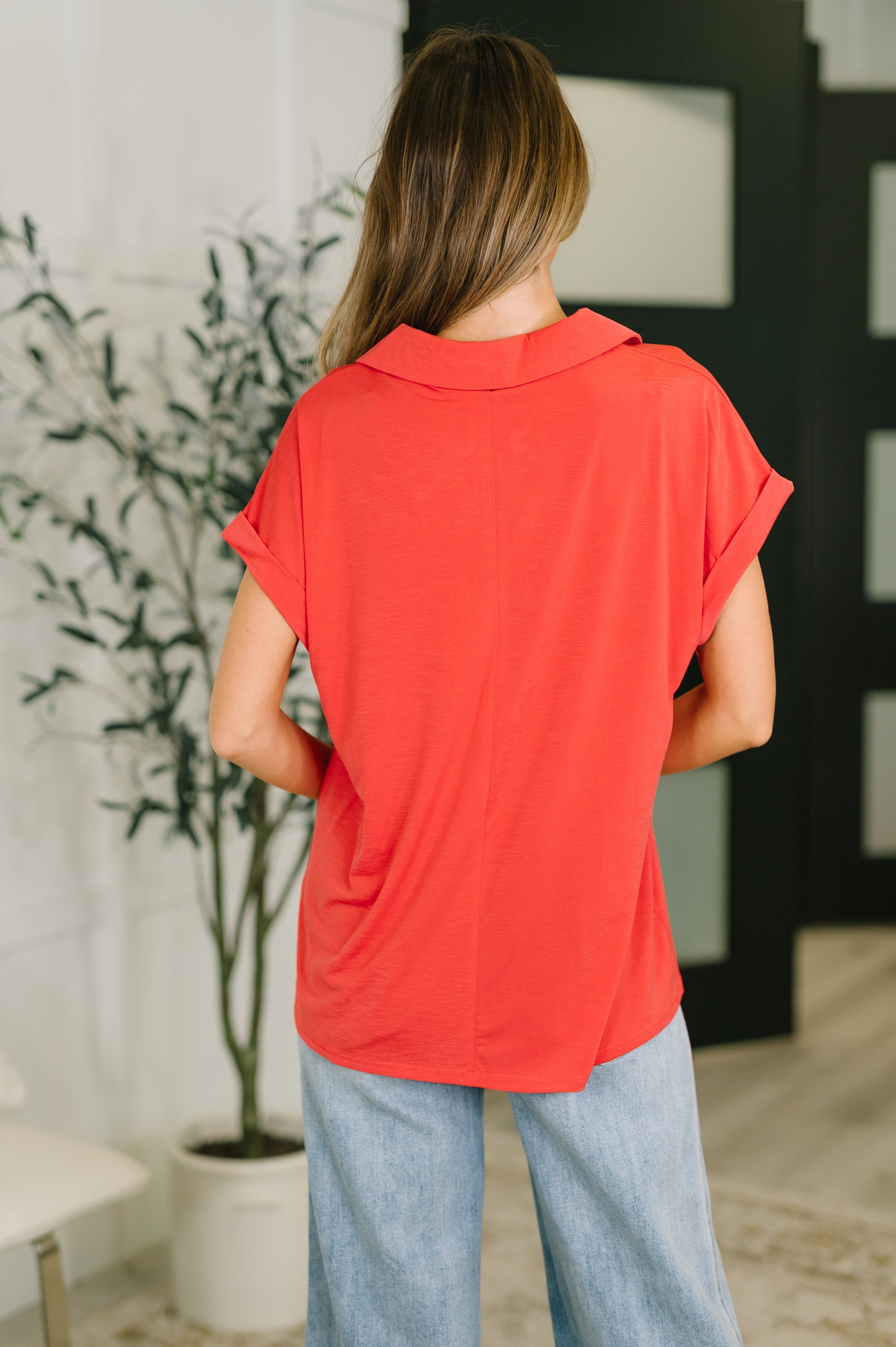 Back view of woman wearing relaxed coral short sleeve collared top with rolled cuffs indoors
