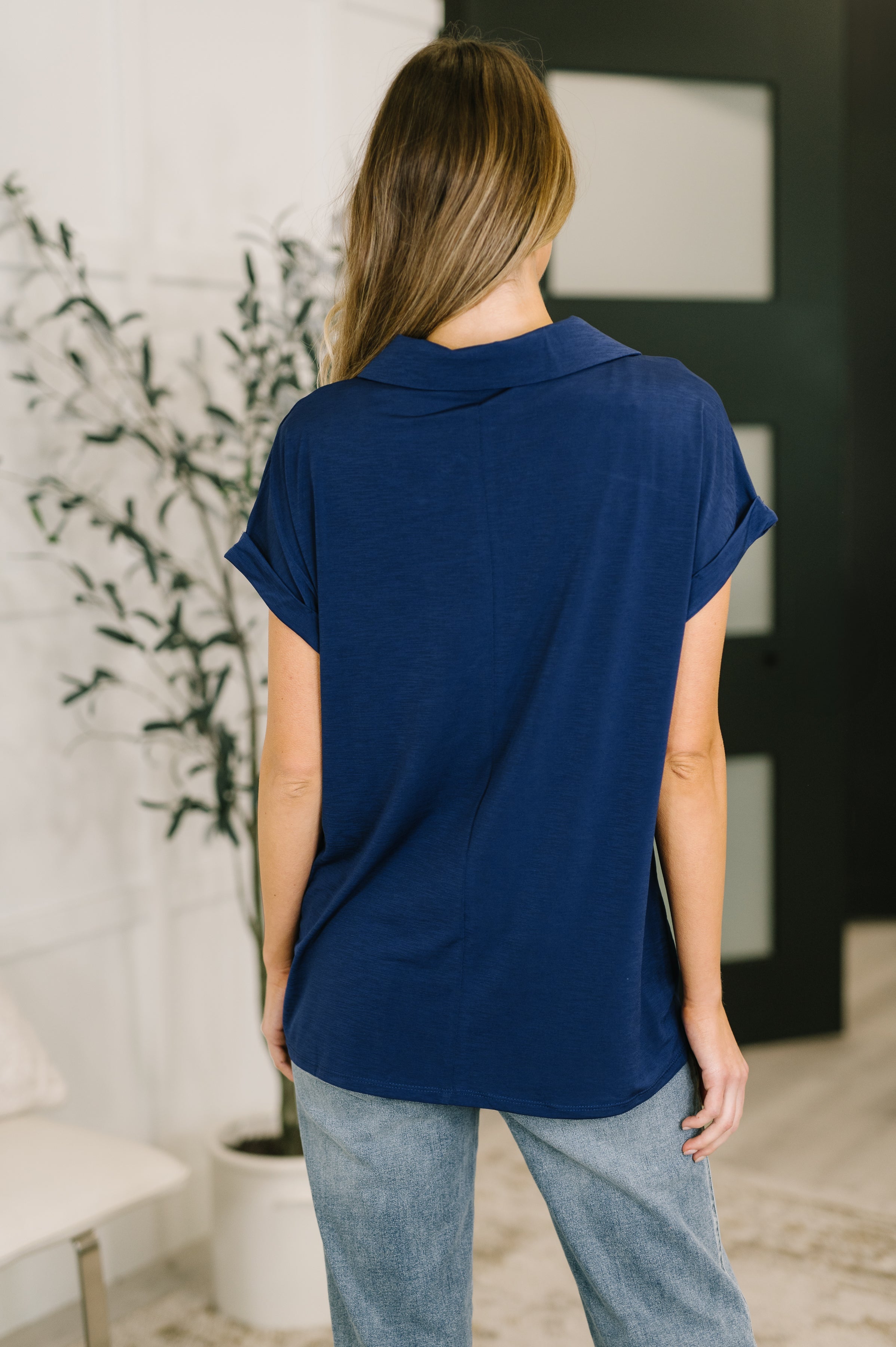 Back view of woman wearing relaxed navy collared top with rolled short sleeves indoors near plant and chair
