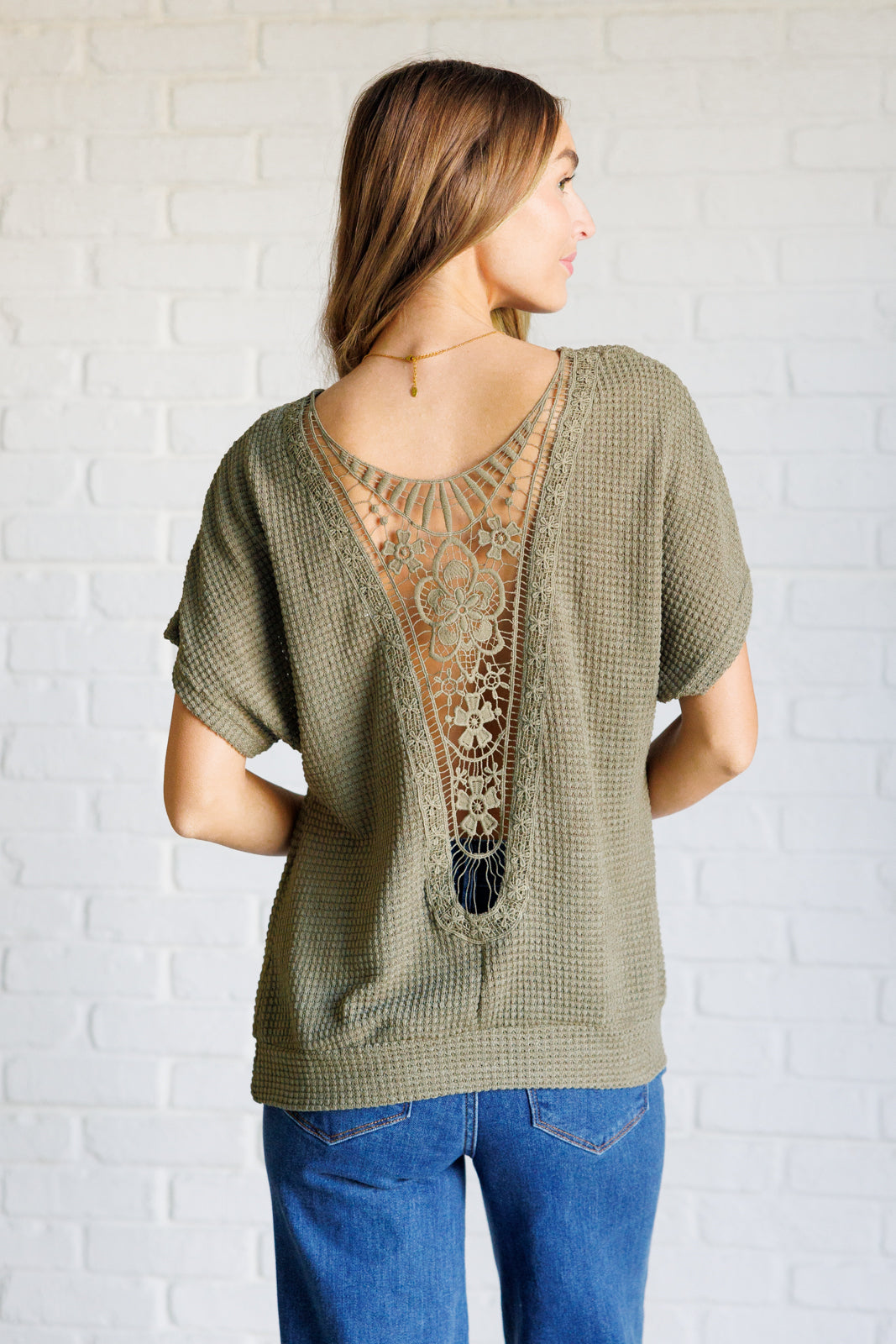 Woman wearing olive waffle knit top with intricate lace back detail paired with blue jeans against white brick wall