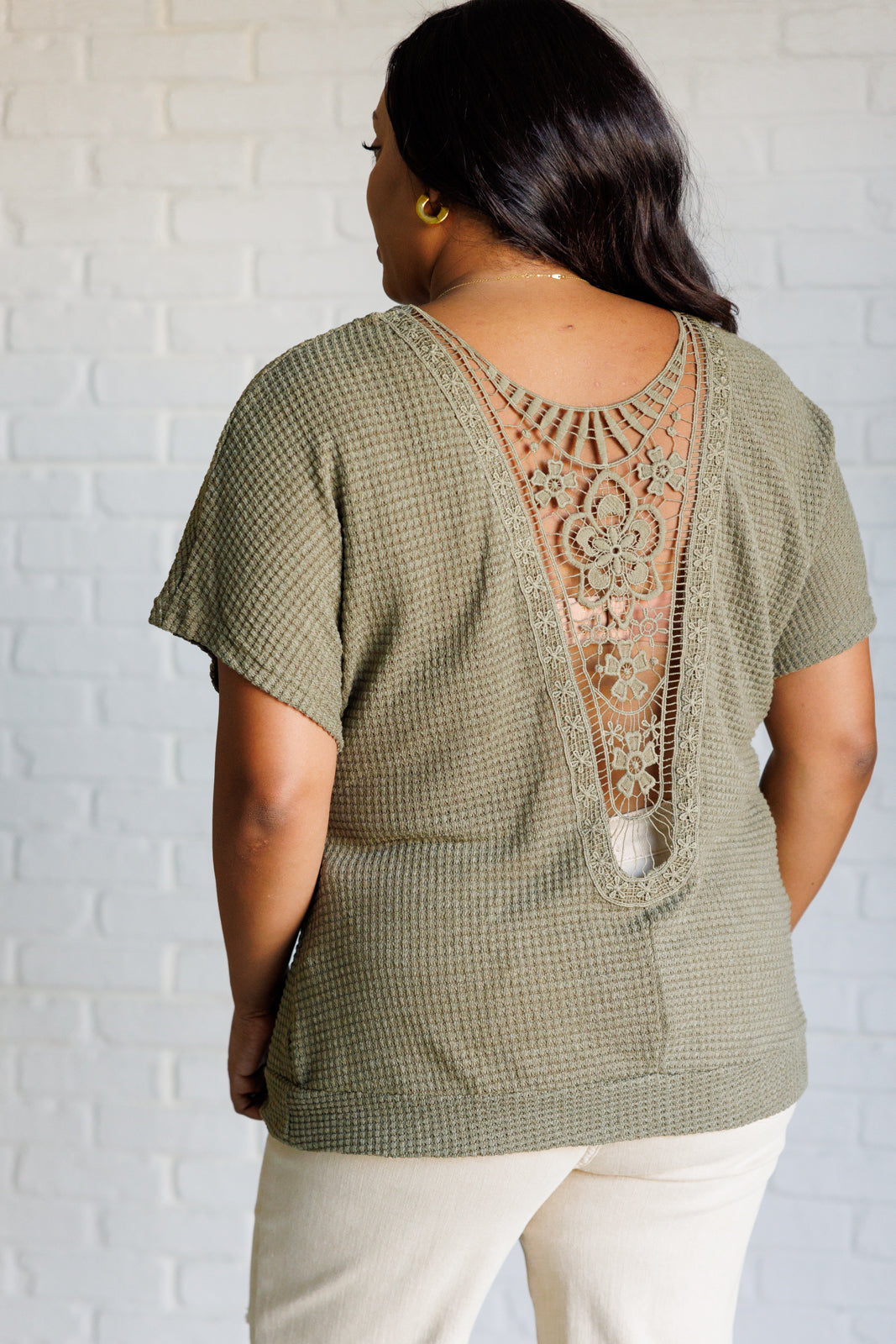 Woman wearing olive textured top with intricate lace back detail paired with light-colored pants standing indoors