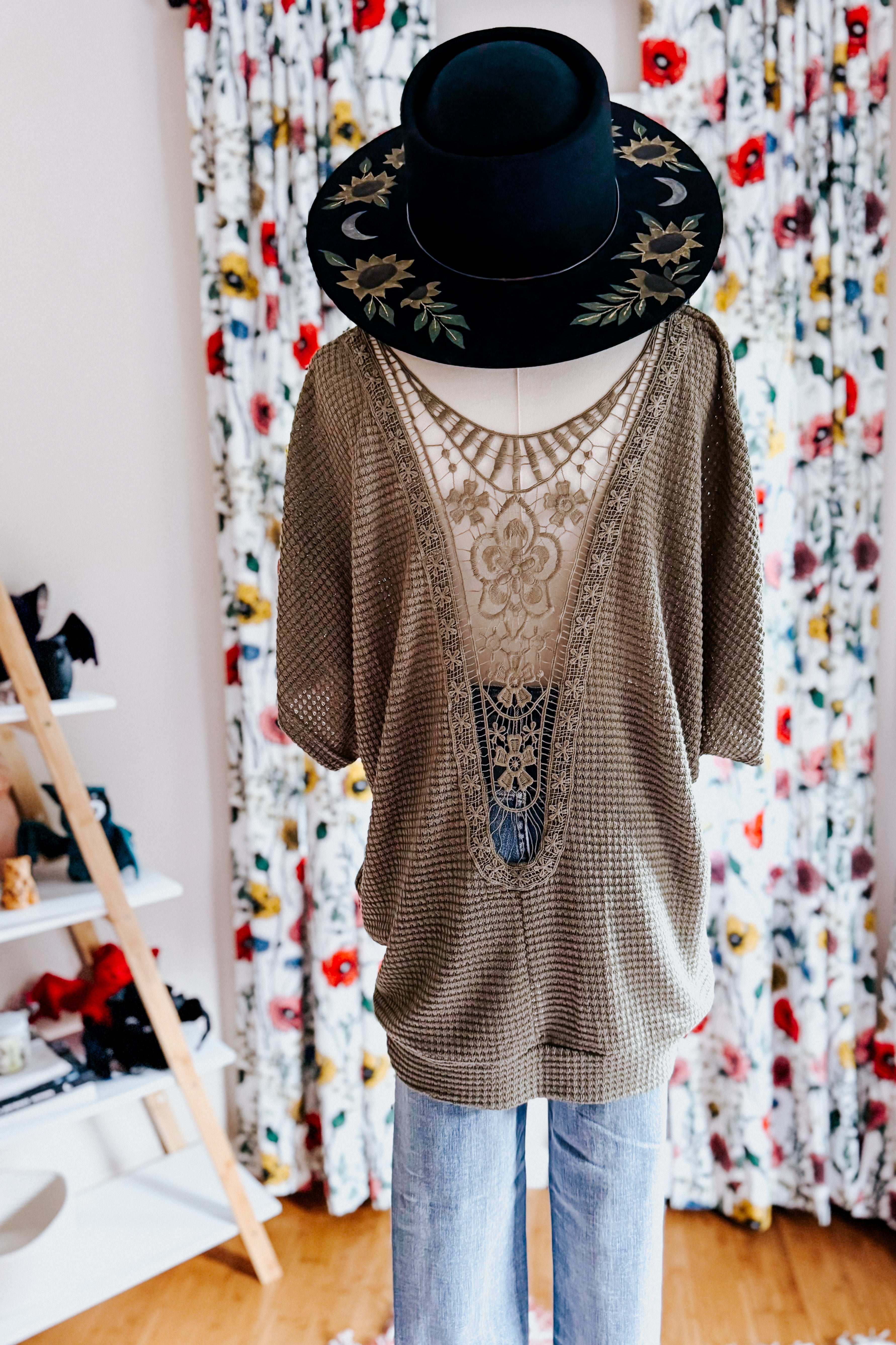 Olive green lace-back top with textured knit and floral embroidery styled with wide-brimmed black hat and light denim jeans