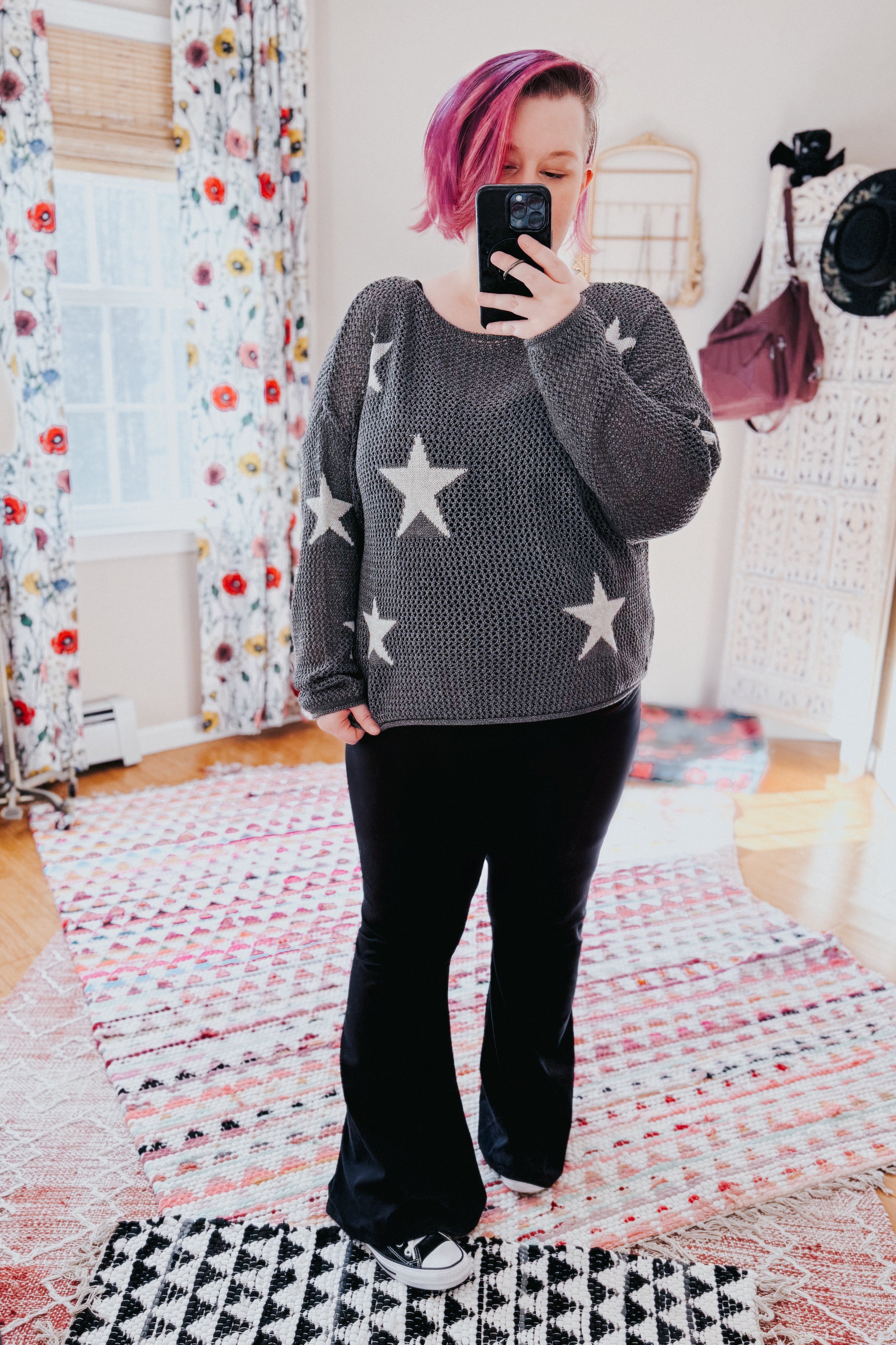 Woman with pink hair wearing charcoal mesh sweater with star pattern paired with black flared pants taking mirror selfie indoors